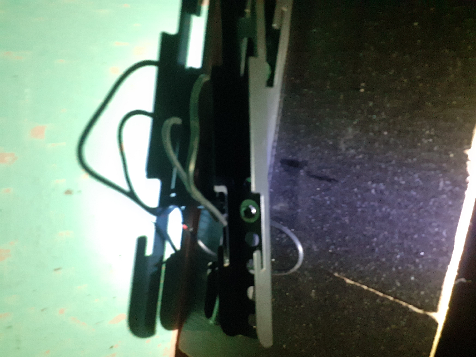 The image shows a close-up, blurry view of a section of a piece of electronic equipment, possibly a printer or scanner, resting on a speckled dark surface that appears to be a desk or countertop. The equipment has a predominantly black and silver color scheme, with some green circular elements visible. The background is a mottled green surface with light-colored splotches, possibly a wall or a textured backdrop. The lighting in the image creates strong shadows and highlights, obscuring some details. There is no visible indication of the location being the city of Jalingo, Nigeria, nor are there any people, actions, or interactions depicted. The scene is static and focused on the object itself.