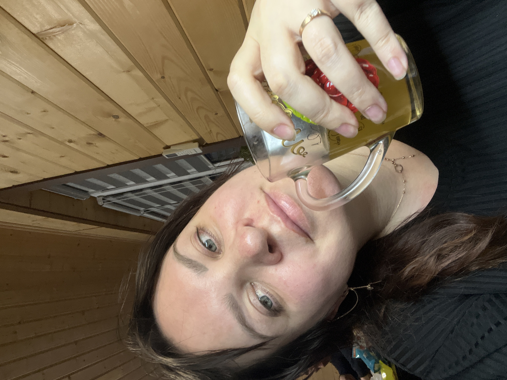 A woman with brown hair and light eyes is holding a glass mug filled with a yellow liquid. She is wearing a ring on her left hand. The background includes a wood-paneled wall and a ceiling fixture. The woman is wearing a dark-colored top and a gold chain. There is no information to suggest that the location is Bol'shakino, Russia.