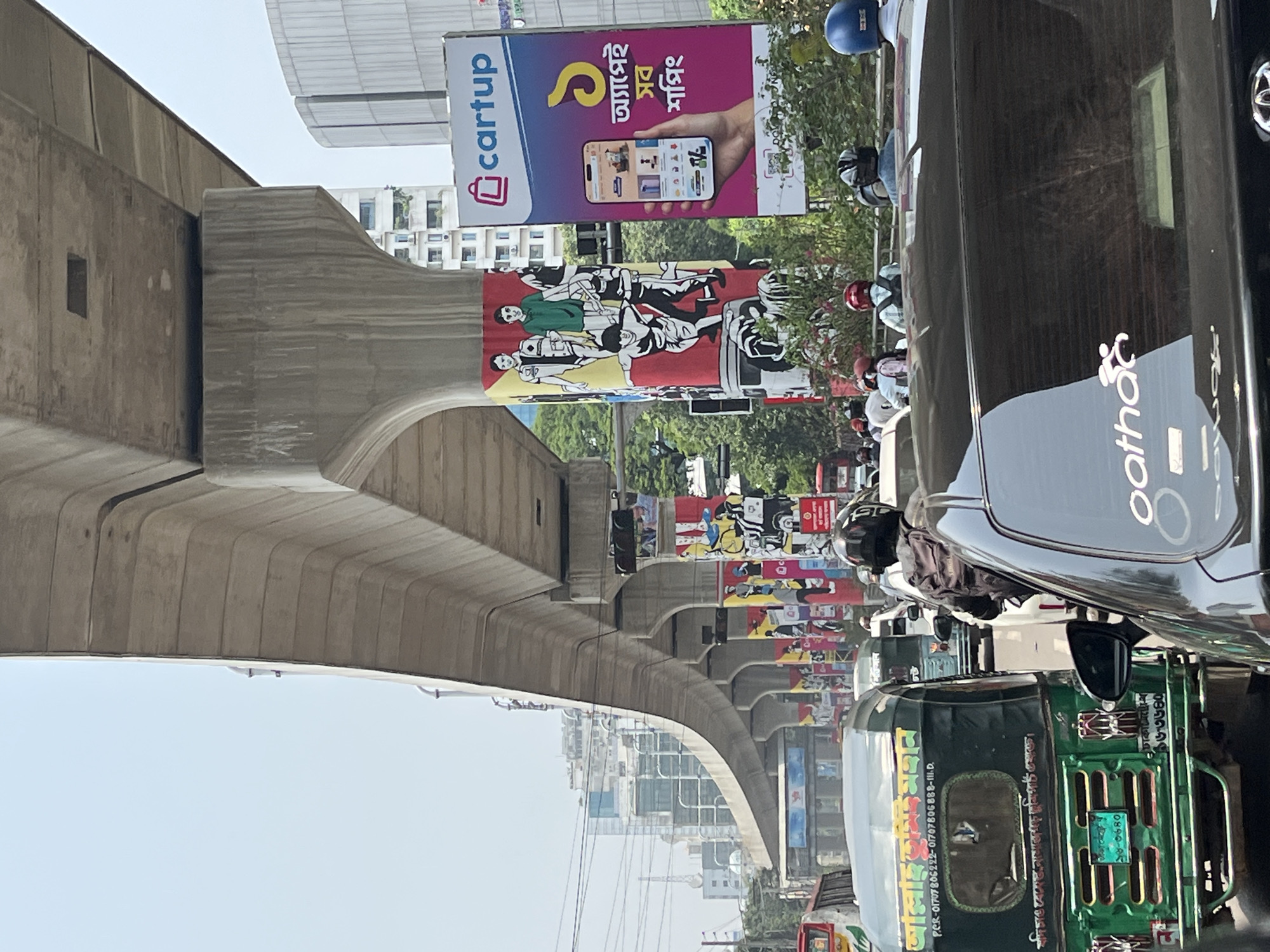 The image shows a busy street scene in Dhaka, Bangladesh, dominated by an elevated concrete structure. The structure, likely an overpass or flyover, curves across the frame, with billboards attached to its side. The billboards advertise "cartup" and feature images and text in Bengali.
Below the structure, the street is crowded with vehicles, including auto-rickshaws (tuk-tuks) and cars. Several people wearing helmets can be seen, likely motorcycle or scooter riders. The "Pathao" logo is visible on the side of a black car. Bengali script and numbers are also clearly visible on the auto-rickshaw, and the phrase "আল্লাহ তুমিই মহান" (Allah Tumii Mohan/Allah you are great) is also visible on the auto-rickshaw. The scene is vibrant and indicative of a bustling urban environment. The presence of the elevated structure and the density of traffic are defining characteristics of the location.