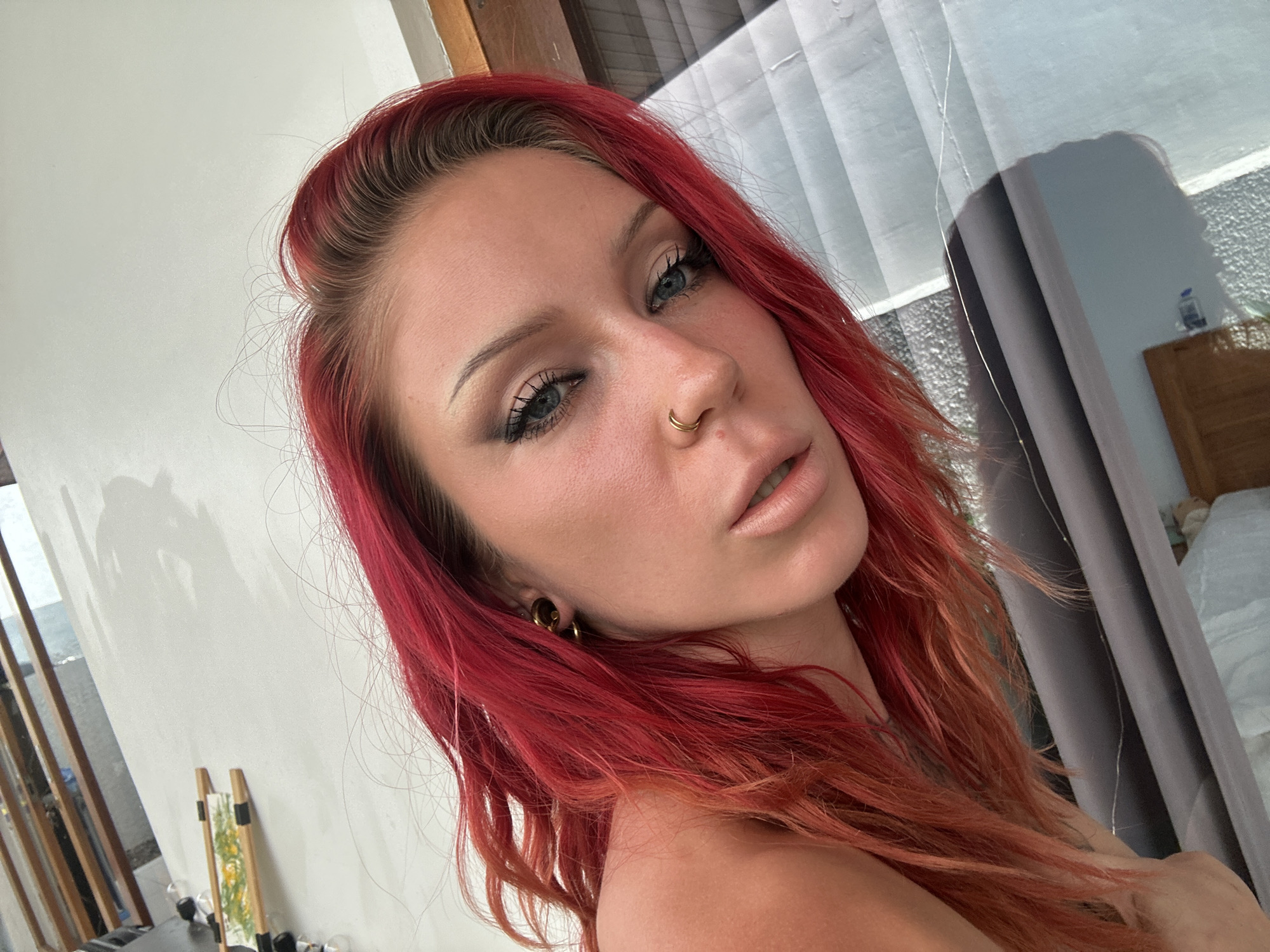 In North Kuta, Indonesia, a selfie is taken by a woman with vibrant red hair. She has blue eyes, prominent eyelashes, and a nose ring. Her lips are slightly parted, and she's wearing light-colored lipstick. Her left ear has a gold earring. The background shows a partially visible room with a window covered by sheer curtains, a wooden wardrobe, and what appears to be a bed. On the wall to the left, there are vertical wooden slats and a white surface with a painting of yellow flowers. The lighting suggests it is daytime.