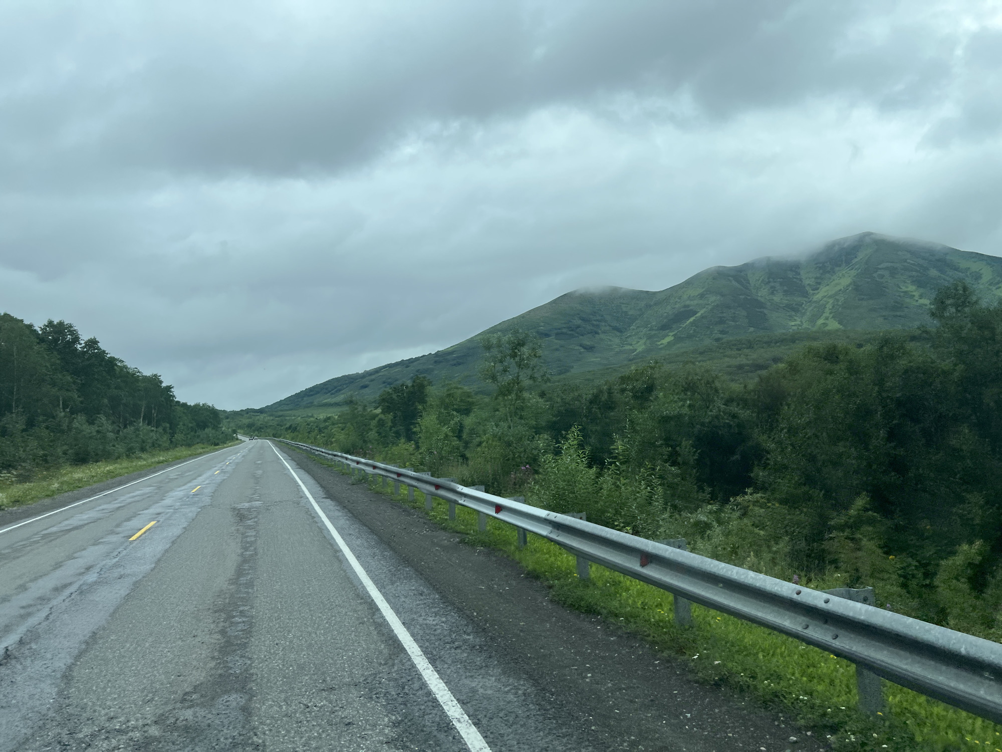 This is a daytime, eye-level, wide-angle photograph taken from a moving vehicle on a paved road in Nachiki, Russia. The road, which appears to be a two-lane highway, is wet and shows signs of wear, with visible patches and yellow and white lane markings. A metal guardrail runs along the right side of the road, with red reflectors attached. The landscape on either side of the road is dominated by lush green trees and rolling hills. In the background, a large mountain range is visible, with its peaks partially obscured by clouds. The sky is overcast with gray clouds. There are no people or animals visible in the image.