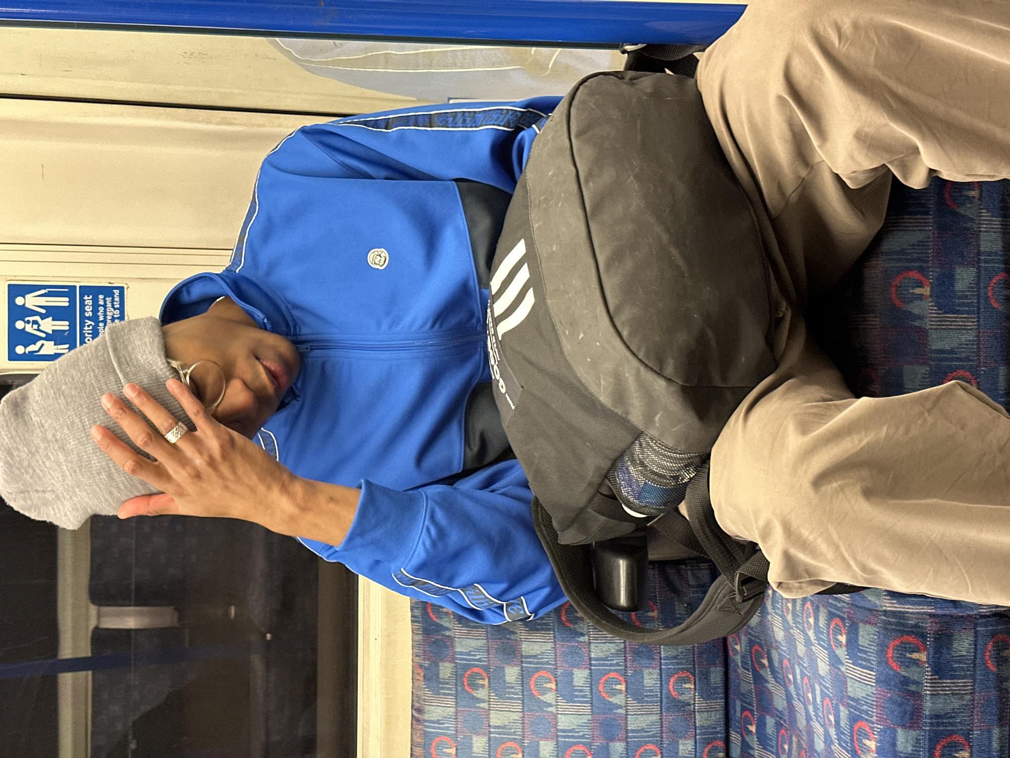 An individual is depicted seated within a London Underground train carriage. The person, appearing male with a medium-to-dark complexion, wears a grey knit beanie, round spectacles, a blue and black Adidas track jacket with white striped details on the sleeves, and light brown trousers. Their right hand, adorned with a silver-toned, patterned ring on the ring finger, is raised to the forehead, touching the beanie. The individual's head is tilted downwards, and their gaze is directed towards a black Adidas backpack with three white stripes resting on their lap. The distinct blue, red, and grey patterned seat upholstery, characteristic of the London Tube, is visible. Above the individual's head, a blue "Priority seat" sign, featuring pictograms of an elderly person, a pregnant person, and an adult with a child, is affixed to a cream-colored wall panel. A dark window is visible on the left, and a blue vertical pole is on the right.