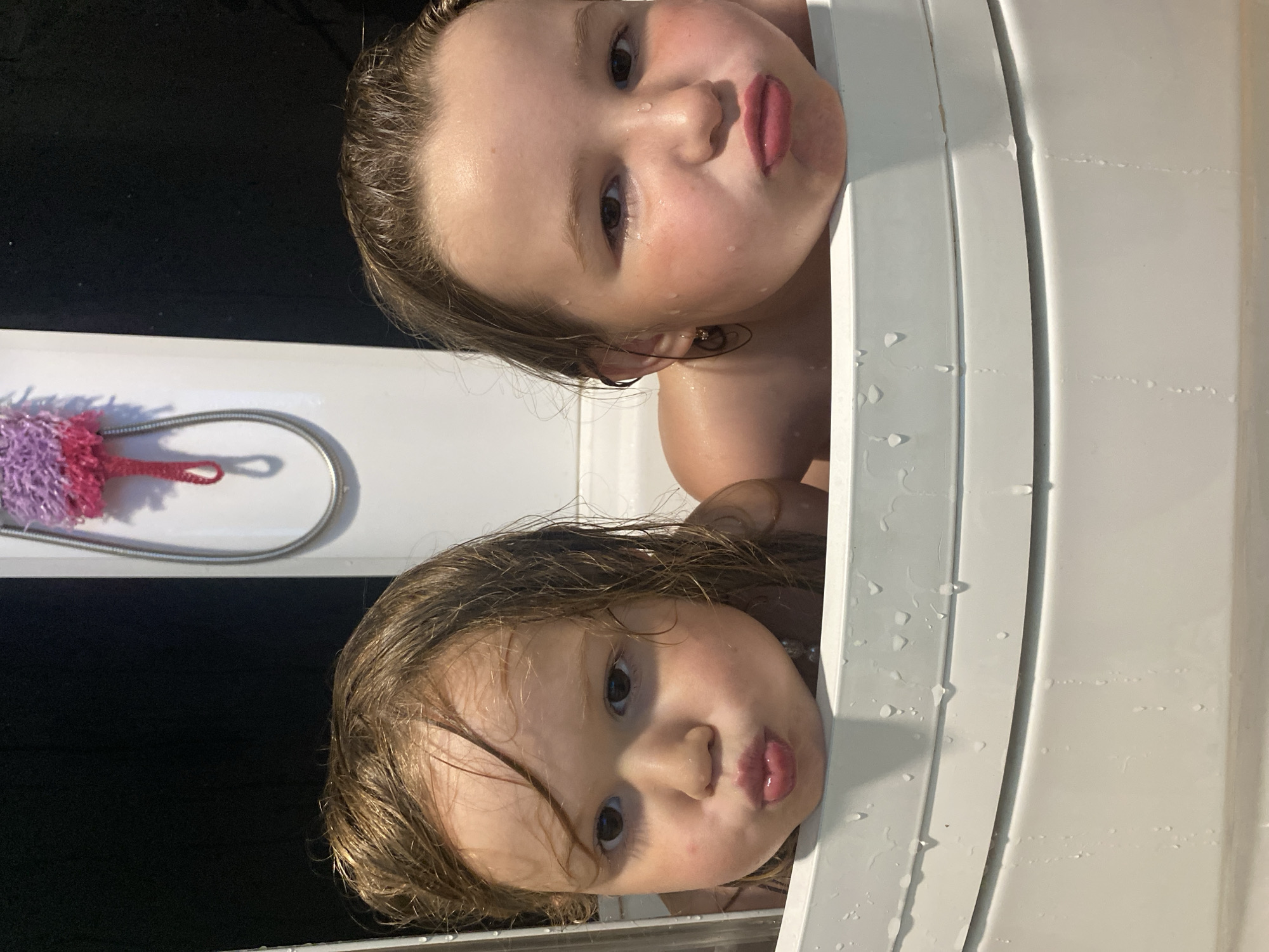 This image, taken in Chaikovsky, Russia, captures a playful moment during bath time. Two young children, likely girls, with wet hair and faces, are seen peeking over the edge of a white bathtub or shower stall. Their faces are oriented sideways, with their heads tilting upwards from the tub.
Both children have wet, light brown hair and fair skin, with visible water droplets on their faces and hair, suggesting they are actively bathing or have just finished. They are making identical expressions, puckering their lips in a "fish face" or a playful kiss, indicating a moment of fun. The child positioned higher in the frame (if rotated, the one on the left) has dark eyes and a slightly rosy complexion. The child below her also has dark eyes and a similar cheerful expression.
A chrome shower hose is visible on the left side of the frame, with a pink and purple bath puff attached. The interior background beyond the tub's edge is dark and out of focus, hinting at a dim bathroom or a dark shower curtain. The scene conveys a sense of candid joy and childhood innocence during an everyday activity. No text is visible in the image.