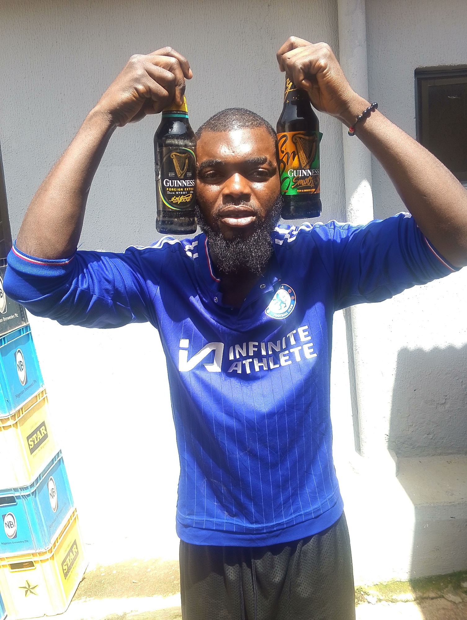 A man with a beard and dark skin is standing outside, holding two bottles of Guinness Stout above his head. He is wearing a blue t-shirt with white stripes on the sleeves and the words "INFINITE ATHLETE" printed on the front. He is also wearing black shorts. Behind him, there are stacks of blue and yellow crates, some labeled "NB Nigerian Breweries Plc" and "STAR". The lighting suggests it is daytime, and the sun is casting shadows, indicating it might be a sunny day. The man's expression is neutral. The scene appears to be in an outdoor setting, possibly a warehouse or a shop.