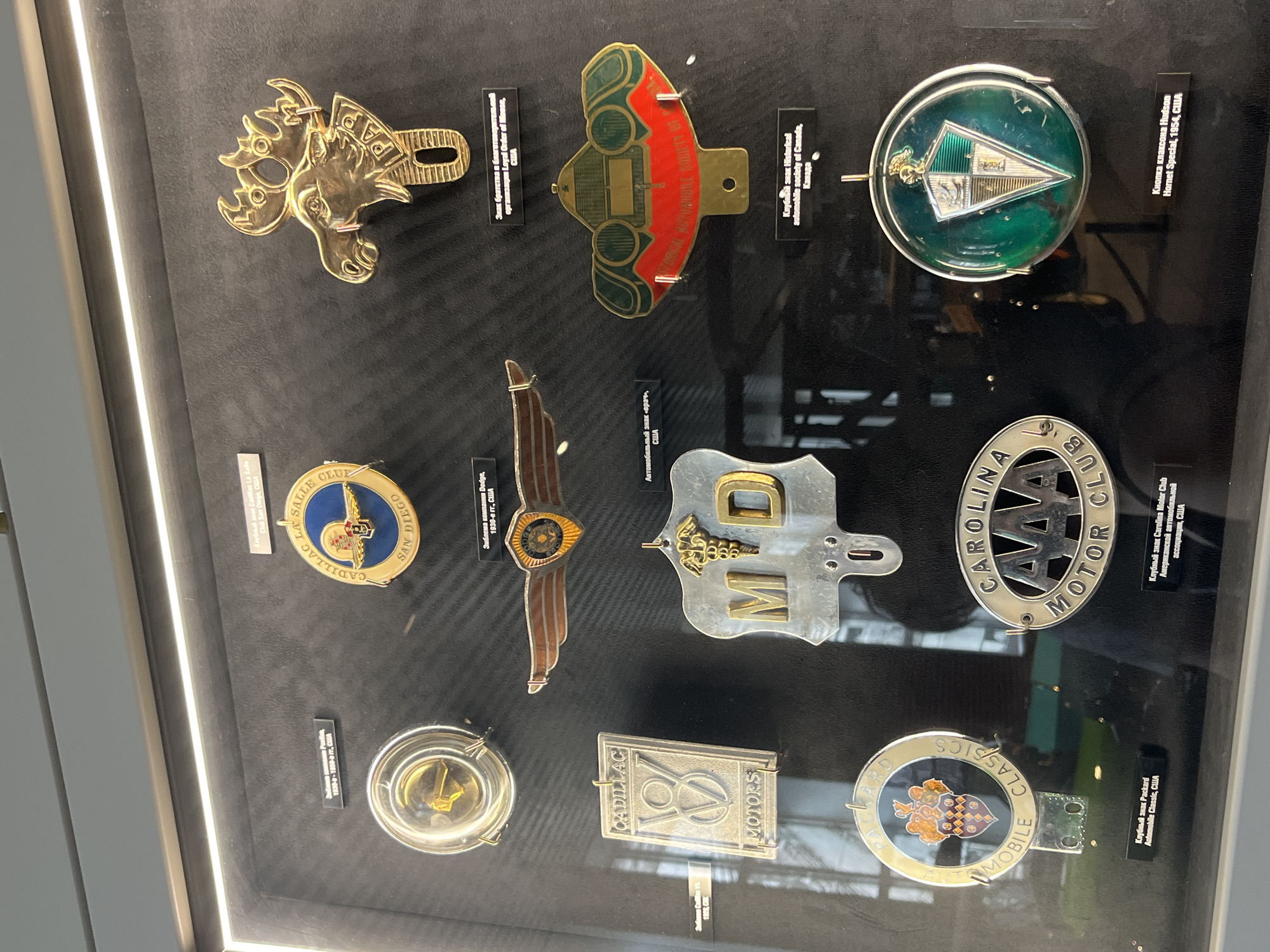 This media file showcases a collection of vintage automobile badges and emblems displayed in a glass case. The objects are arranged in rows on a dark background. Visible text on the labels indicates they are from various automobile clubs and historical societies, including "Cadillac La Salle Club San Diego," "Historical Automobile Society of Canada," and "Carolina Motor Club." Some of the emblems themselves have visible text such as "CADILLAC," "MOTORS," and "CLASSICS." There are also specific vehicle models mentioned like "Hudson Hornet Special." The lighting suggests an indoor setting, likely a museum or exhibition, with bright lights illuminating the display. The collection appears to be a historical exhibit dedicated to automotive memorabilia. The location context provided is Verkhnyaya Pyshma, Russia, which might be the location of this display. No people are visible in the media file, and the focus is entirely on the objects.