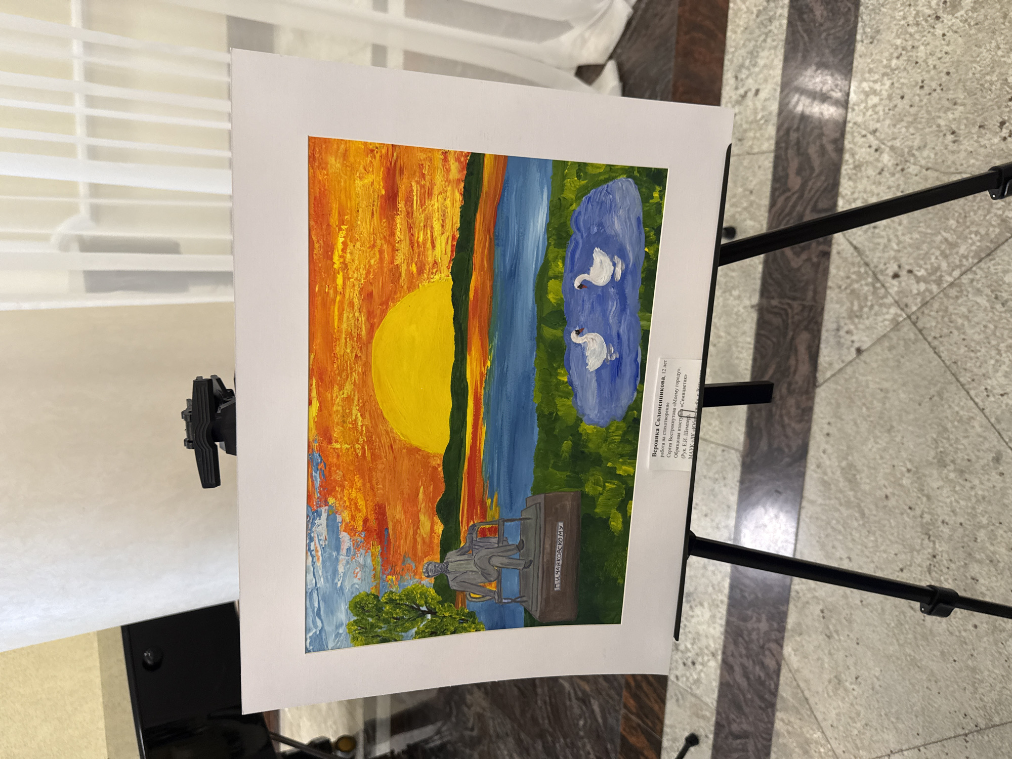 The image shows a child's painting displayed on a stand. The painting features a scene with a large orange sun setting (or rising) over a landscape with a river, trees, swans in a pond, and a statue of a man sitting on a bench. The name "П.И.ЧАЙКОВСКОМУ" is inscribed on the bench.
A label attached to the painting identifies the artist as Вероника Соломенникова, a 12-year-old from Votkinsk, Russia, who created the artwork based on a poem by Сергея Вострокнутова titled "Моему городу" ("My City"). The painting was created as part of a studio project called "Семицветик" under the guidance of E.I. Шемпер. The studio is associated with the Municipal Autonomous Cultural Institution (МАУК) "ЛК "Юбилейный," which is located in Votkinsk. The stand is set against a light-colored wall and a floor with dark and light stripes.