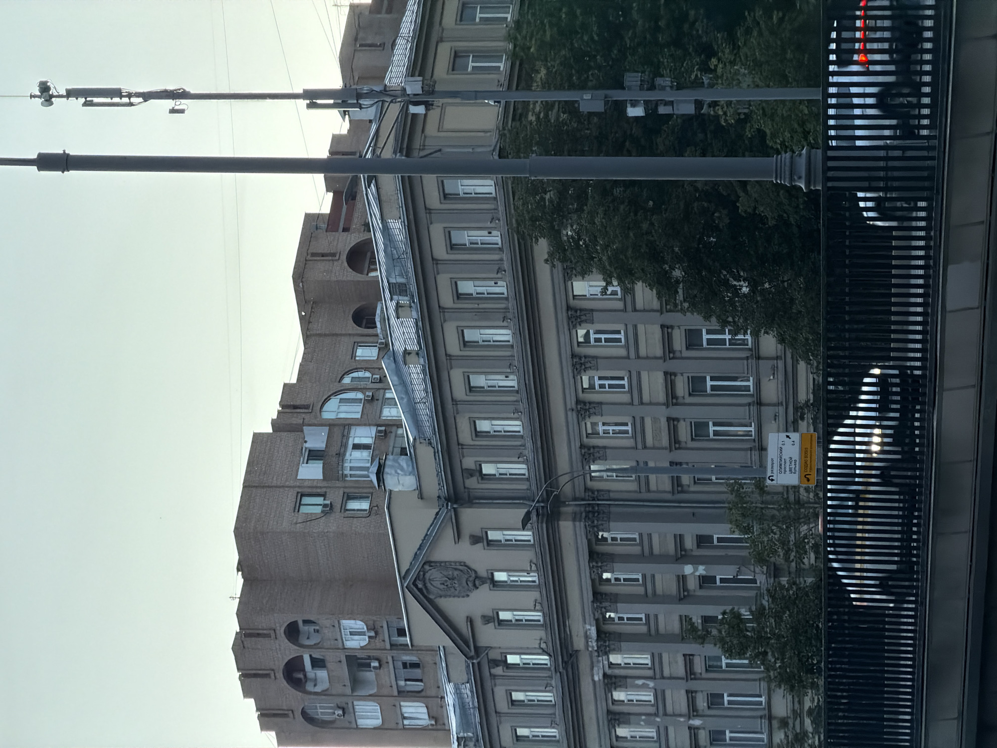 The image captures an urban street scene in Moscow, Russia, under a clear, bright daytime sky. In the foreground, a black metal railing of a bridge or overpass runs along the right side, with faint reflections of vehicle lights visible below. Two utility poles are central: a thicker dark grey one and a taller, slender pole fitted with what appear to be surveillance cameras or communication equipment near its top.
The background features several multi-story buildings. On the left, a brick building exhibits multiple levels with distinctive arched windows and balconies, some of which are enclosed. To its right, a more ornate, lighter-colored building with classical architectural elements, including cornices and decorative facade details, dominates the middle ground. Further right, another similar building is partially visible. Green foliage from trees is interspersed among the structures and behind the utility poles.
A white rectangular road sign, written in Cyrillic, is visible on one of the poles. It provides navigational directions, listing "U-turn" (разворот) and distances to several Moscow streets: "Olimpiyskiy Prospekt" (Олимпийский проспект) at 1.1 units, "Butyrskiy Val" (Бутырский вал) at 0.4 units, "Sushchevskiy Val" (Сущёвский вал), and "Ulitsa Pravdy" (УЛ. ПРАВДЫ). An advertisement below these reads "ПОСАДКА BOGUS" (Bogus Landing/Boarding), with "ТРОТУАР" (Sidewalk) underneath, possibly indicating a transport service pick-up point. No people are visible, suggesting a moment of urban tranquility.