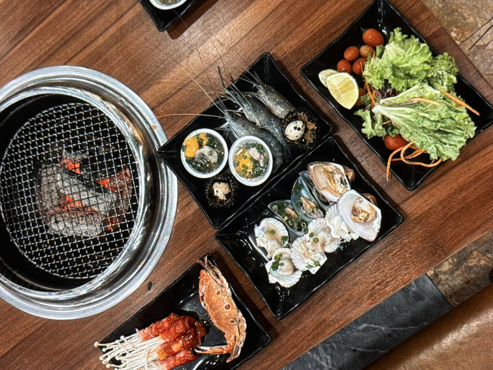 The image depicts a table setting for a seafood BBQ meal, likely in a restaurant in Đà Nẵng, Vietnam. A circular silver metal grill sits to the left, with visible charcoal and glowing embers. Four black square plates are arranged around the grill. One plate holds raw shrimp, two bowls containing egg yolk and greens, and sea urchin. Another plate has an assortment of oysters, clams, and scallops. A third plate contains a salad with lettuce, cherry tomatoes, lemon wedges, and shredded carrots. The final plate displays enoki mushrooms wrapped in meat, and a grilled crab. The table is made of wood with a dark stain. A light-colored stone-like surface is visible in the bottom right corner. The scene suggests a casual dining experience focused on grilling fresh seafood.