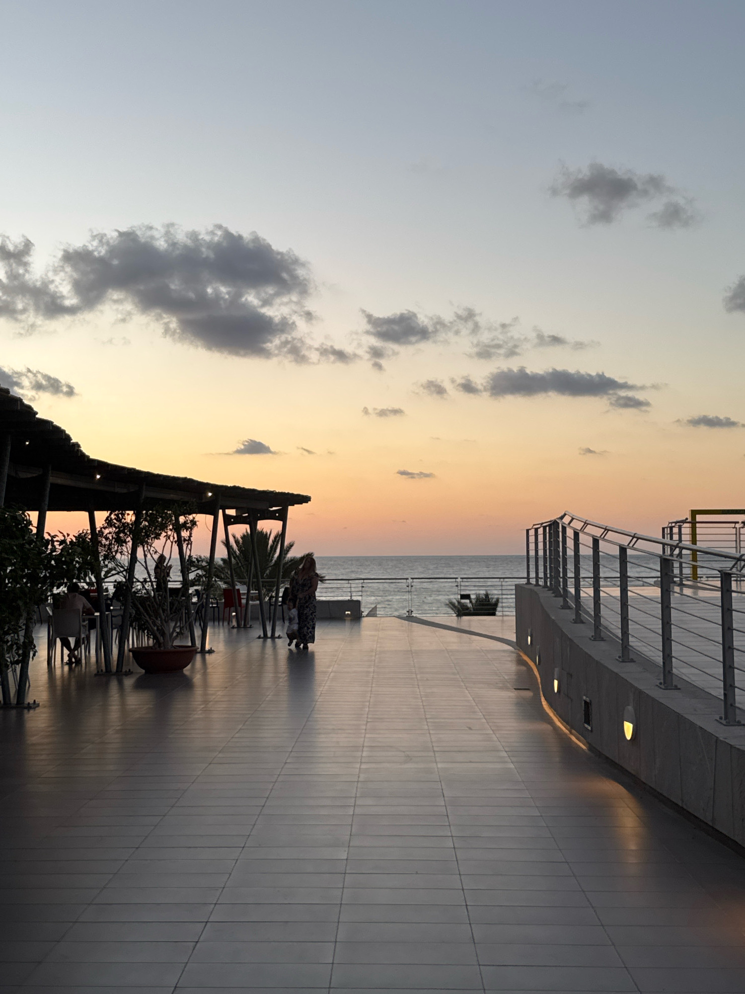 The image depicts a seaside promenade at dusk in Isola delle Femmine, Italy. The sky displays a gradient of pale blue at the top, transitioning to soft orange and yellow hues near the horizon, indicating the setting sun. Scattered clouds, some dark grey and others lighter, are visible in the upper portion of the sky.
In the foreground, a wide, tiled promenade stretches into the distance. To the left, an outdoor seating area with tables and chairs is partially covered by a dark, sloped roof supported by pillars. Trees and potted plants are arranged around the seating area, with some patrons visible sitting at the tables. A woman and a child are walking away from the camera on the promenade, towards the sea. The woman is wearing a long, patterned dress, and the child is smaller.
To the right of the promenade, a curved barrier with a metal railing runs along the edge. The barrier is constructed of stone and concrete, with several illuminated wall lights casting a warm glow onto the promenade. The sea is visible beyond the railing, with a calm surface reflecting the twilight sky. In the distance, a few palm trees can be seen near the treeline. The overall atmosphere is serene and tranquil, characteristic of an evening by the sea.