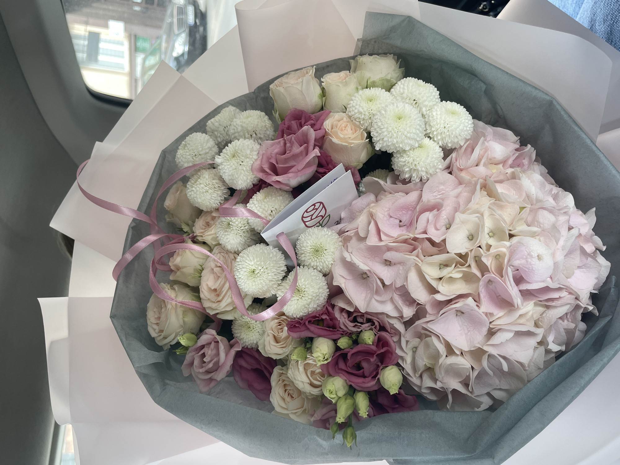 The image shows a bouquet of flowers with pink and white roses, white chrysanthemums, purple eustomas, and a large pink hydrangea bloom. The bouquet is wrapped in grey and white paper and tied with a pink ribbon. A small white card with a red logo is tucked into the bouquet. The bouquet is inside a car, as indicated by the car window and interior visible in the background. The location appears to be urban, with buildings seen through the car window. The city and specific location are unknown.