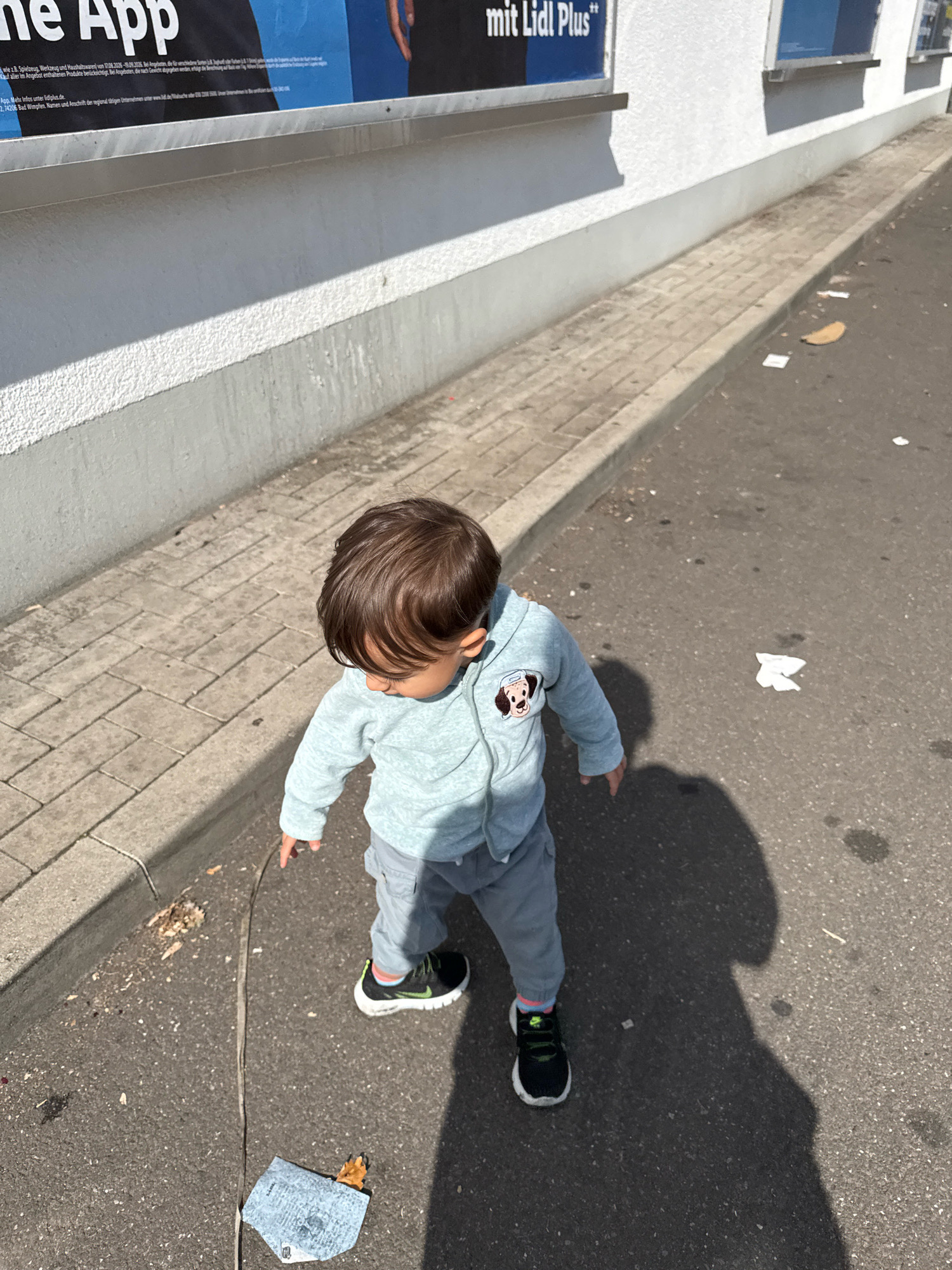 The image shows a young child, likely a toddler, walking on an asphalt surface next to a sidewalk. The child is wearing a light blue hooded jacket with a cartoon dog patch on the chest, light gray pants, and black sneakers with white soles. The child is looking down and appears to be reaching towards something on the ground. The environment is an urban or semi-urban setting, with a building wall in the background. There is some debris on the ground, including what appears to be a crumpled piece of paper and a fallen leaf. The sun is casting a strong shadow of the child onto the asphalt. Based on the provided information, the location is Überherrn, Germany.