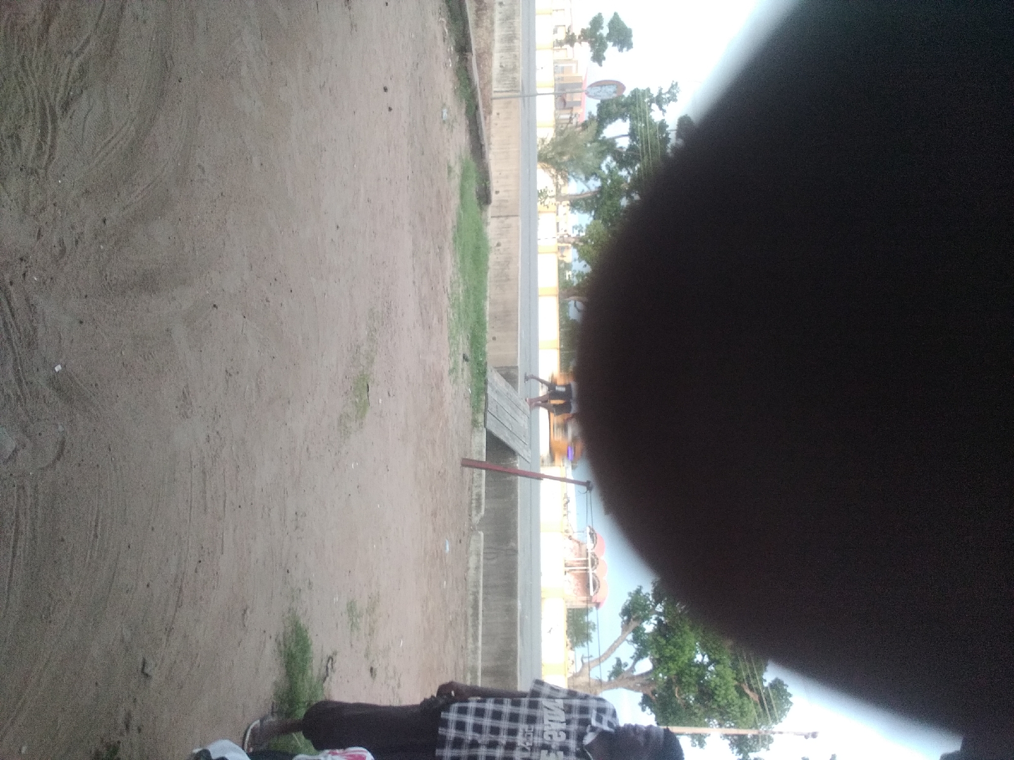 The image depicts a street scene in Jalingo, Nigeria. In the foreground, a person stands on a sandy surface. The person is wearing a black and white checkered shirt and dark pants. The background features a road with traffic, trees, and buildings. There is a speed bump on the road, and a wooden structure is also visible. The sky is overcast, and the overall lighting suggests it is daytime. The scene appears to be an outdoor public space, possibly near a market or a busy intersection. The image is partly obscured by a large dark blur at the top, likely caused by the camera lens or a hand.