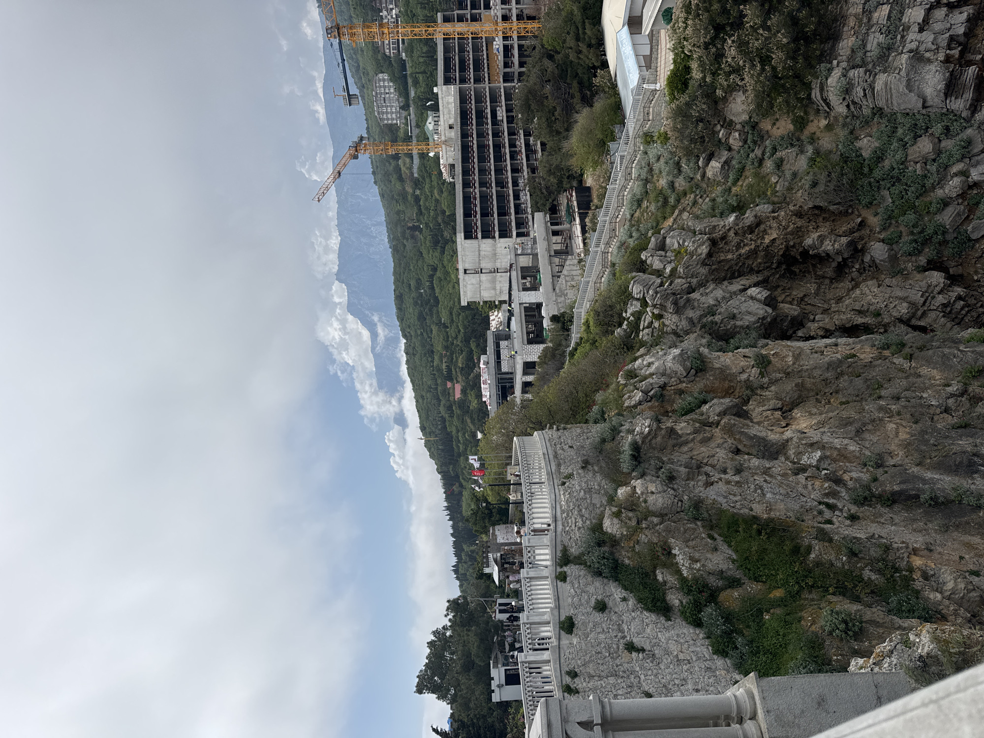 The image shows a landscape with buildings under construction, a rocky cliff, and a cloudy sky. Two yellow cranes stand next to a high-rise building skeleton under construction. Below, buildings with white railings are visible. The foreground features a stone wall and balcony. The location appears to be in the Гаспринский поселковый совет, Ukraine.