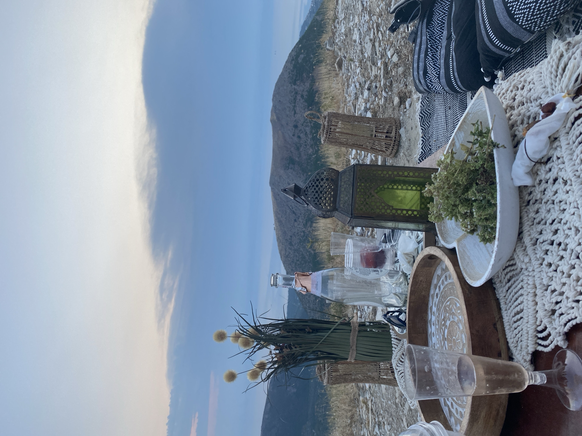 A picnic is set up on a rocky, dry hillside overlooking a valley and distant mountains under a cloudy sky. In the foreground, a champagne flute filled with a pale pink beverage sits on a macrame placemat. To the right of the placemat is a white bowl containing green herbs. A wooden serving tray with an engraved circular pattern is placed behind the champagne flute. A bottle of champagne, two empty glasses, and a small container with a red item are arranged behind the tray. A green glass lantern sits to the right of the champagne bottle. A woven cylindrical basket is positioned to the left of the champagne bottle, and another identical basket is to the right of the lantern. A bunch of dried floral stems is in a green vase on the left side of the scene. In the background, rolling hills are visible, with a larger mountain in the middle ground. The overall scene suggests an outdoor dining setting, possibly at dusk, in a natural, elevated environment. The specific location in Prinos, Greece is not visually identifiable from the image alone.
