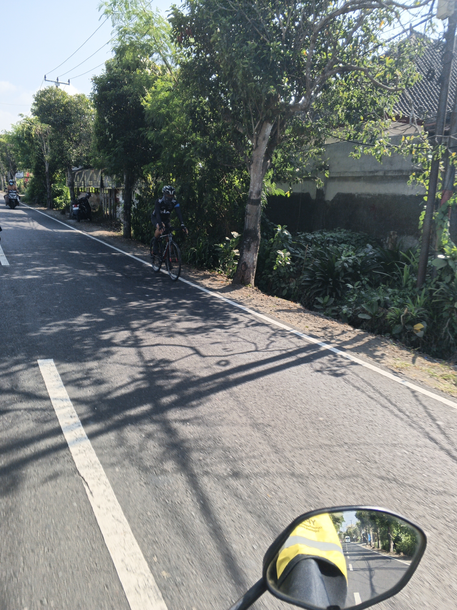 The image depicts a roadside scene in Abiansemal, Indonesia. A cyclist in black attire is riding on the asphalt road, casting a shadow on the surface. The road has a white lane marker. To the left, a motorcycle is parked, and another motorcycle is moving away. A person wearing a blue shirt is on the moving motorcycle. Trees line the road, with dense foliage and a few visible buildings with tiled roofs in the background. The sky is clear and blue. The scene is captured from the perspective of a motorcycle, with its side mirror visible in the foreground, reflecting the road and the cyclist. A person wearing a yellow vest is partially visible within the mirror's reflection.