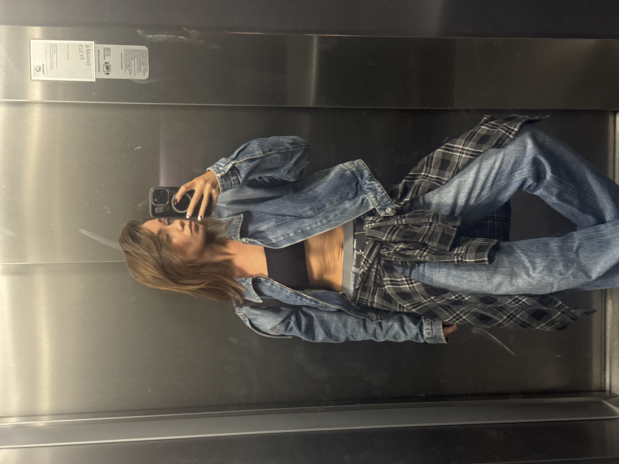 A woman in a black top, denim jacket, plaid shirt tied around her waist, and jeans is taking a selfie in an elevator. The elevator's interior is stainless steel with two informational stickers on the upper left side. The lighting is even, and the reflection on the metal surface adds depth. There is no direct indication of the location being in Budva, Montenegro.