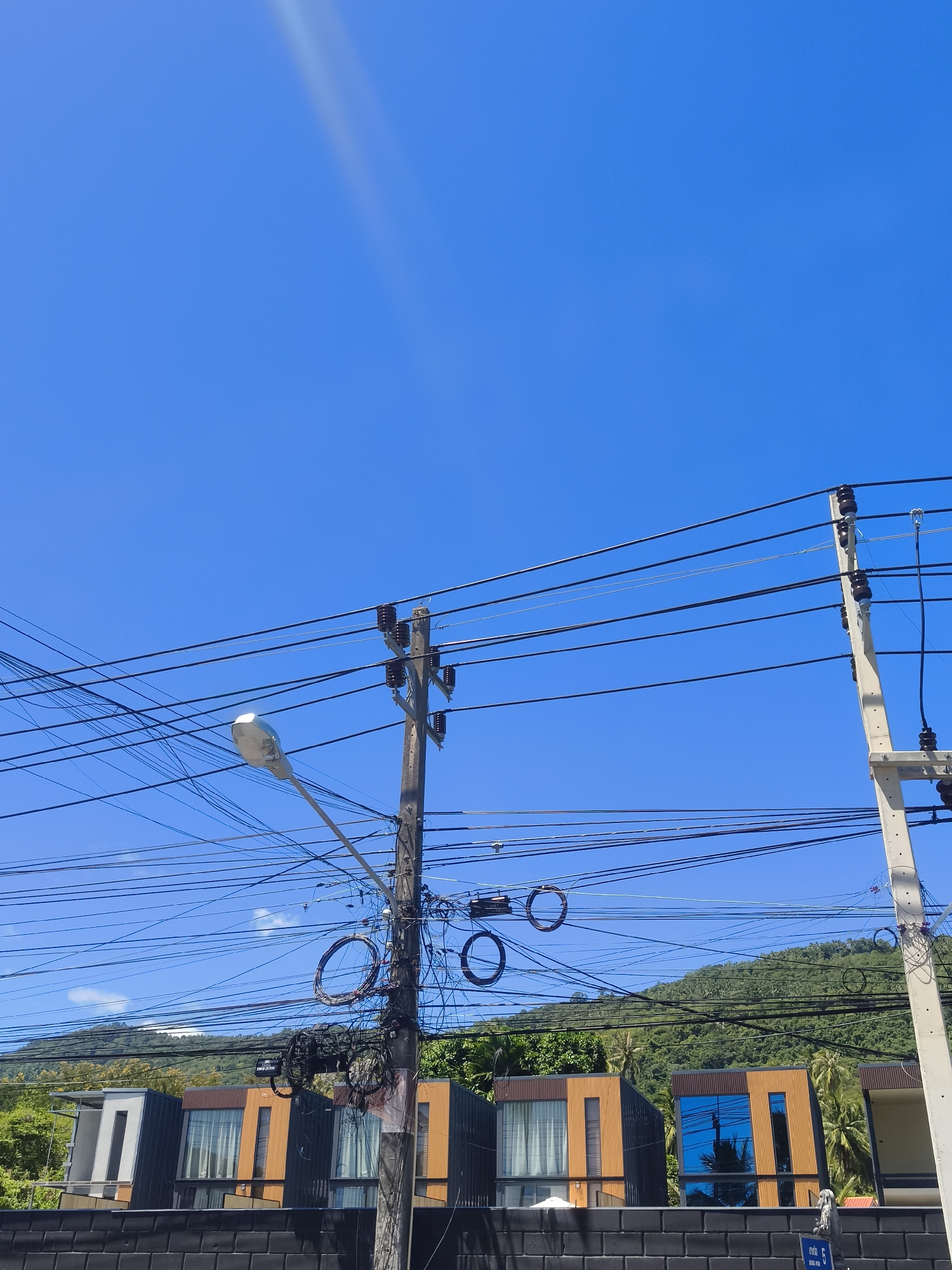 This is a full shot, eye-level, outdoor, daytime, vertical photograph of a street scene in Baan Thong Po, Thailand. The clear, bright blue sky dominates the upper portion of the image. In the foreground, a cluster of utility poles with numerous tangled black electrical wires spans across the frame, creating a dense web of lines against the sky. One pole features a street lamp. The poles are concrete with some wooden elements.
Below the wires, modern, multi-story buildings with dark grey and orange facades are visible. Some of these buildings have large windows, some of which reflect the blue sky and palm trees. A dark, textured wall runs along the bottom of the frame in front of the buildings. In the background, a lush, green, tree-covered hill rises under the blue sky. The overall impression is of a tropical urban environment with a heavy concentration of overhead electrical infrastructure. There are no people or animals visible in the image.