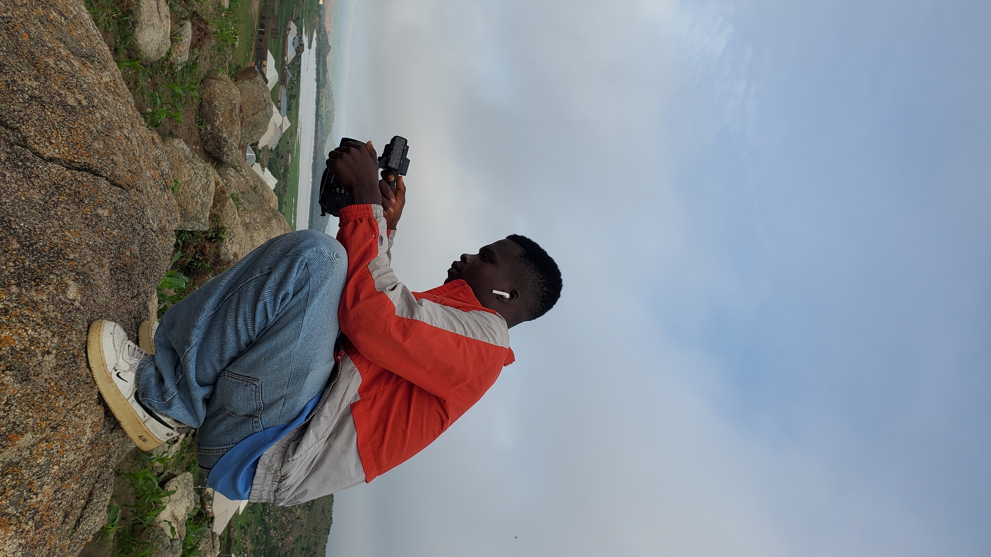 A young Black male, wearing a red and grey jacket, blue jeans, and white sneakers, is crouched on a rocky outcrop. He is holding a camera and appears to be looking at it. He has white earbuds in his ears. In the background, there is a body of water, some buildings, and rolling hills. The sky is overcast. This scene is in Jos, Nigeria, specifically in the Bukuru area, known for its rocky landscape and surrounding villages.
