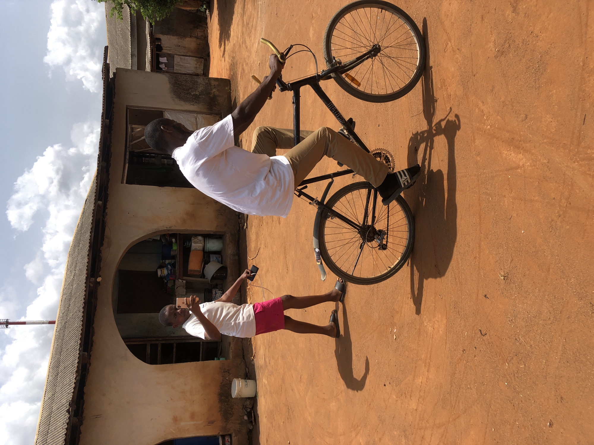 A medium shot depicts two men and a bicycle in Unknown, Unknown. One man balances on a bicycle with gold handlebars wearing a white t-shirt, tan pants, and black Adidas sneakers. The other man wears a white t-shirt, red shorts, and black sandals, using a smartphone. They are on a dirt surface near a weathered building with an arched entrance and a corrugated metal roof. A tall communications tower is visible in the sky.