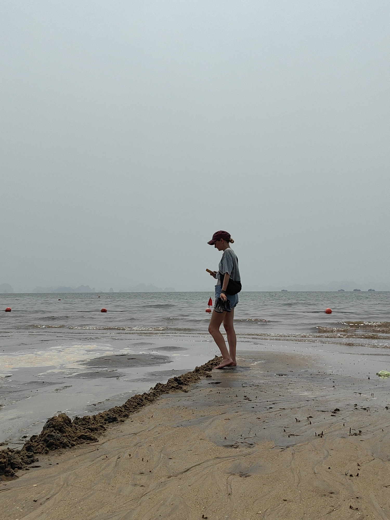 A woman stands on the beach at Bai Chay Ward, Vietnam, on a cloudy day. She is wearing a burgundy baseball cap, a grey t-shirt, and denim shorts, and she is looking at a mobile phone in her hand. A black shoulder bag is across her chest. The beach is sandy with some dark patches. The water has some small waves and there are orange buoys visible in the distance. Hazy islands are visible on the horizon.