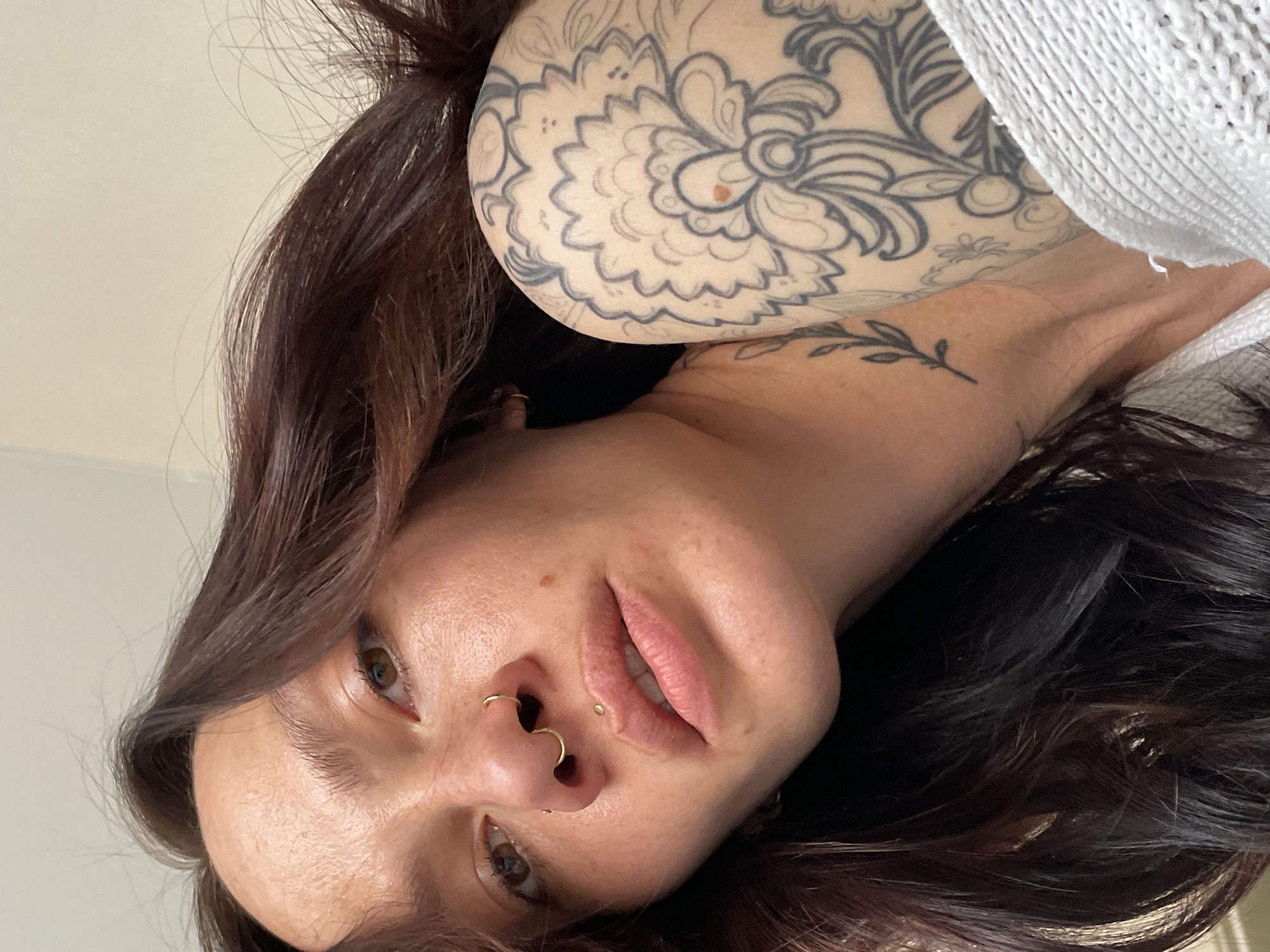 A woman with brunette hair and various tattoos poses from an overhead angle. She has fair skin and is wearing two gold-colored nose rings. One tattoo is of a floral design on her shoulder and upper arm, while a smaller tattoo of leaves is on her wrist/forearm area. She is wearing a white, loosely knit garment. The background is a plain, light-colored wall. There is no evidence suggesting a location in Izhevsk, Russia.