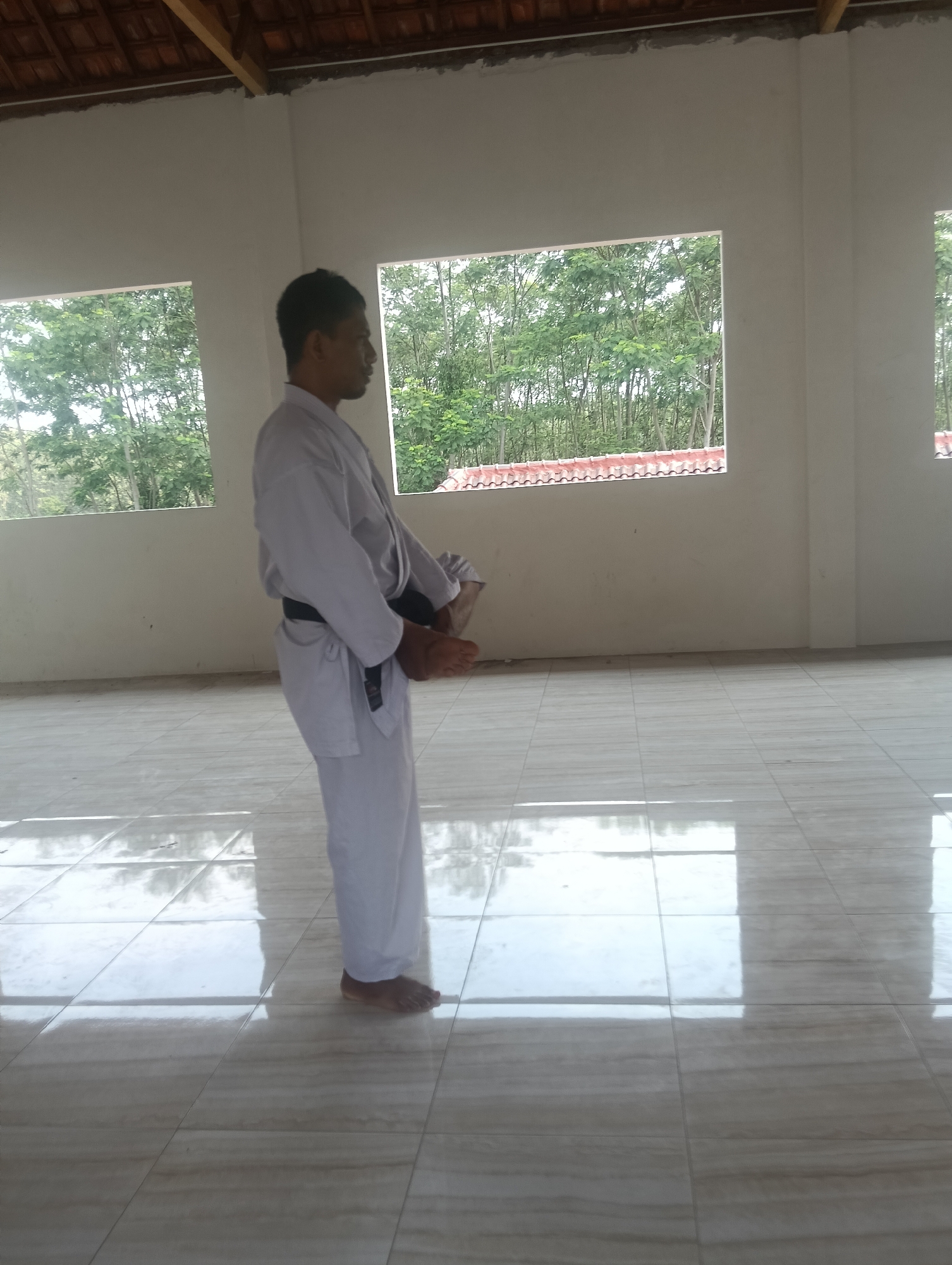 A person wearing a white karate gi with a black belt stands in a room with tiled flooring and large windows. The windows offer a view of lush green trees. The room appears to be unfinished, with exposed wooden beams on the ceiling. The person is in a martial arts stance, with their arms positioned in front of their body and their right foot lifted. The lighting suggests it is daytime. The location is identified as Adinuso, Indonesia.