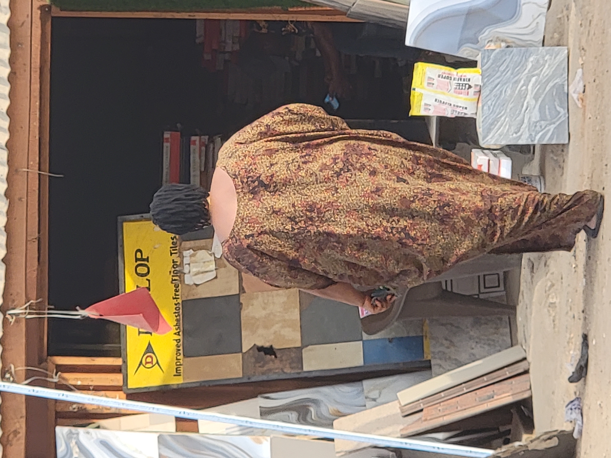 A woman in a patterned brown and tan dress is bent over looking at tiles on the ground outside a store in Maiburiji, Nigeria. The store displays a Dunlop sign with "Improved Asbestos-Free Floor Tiles" written on it. Ceramic tiles are for sale.