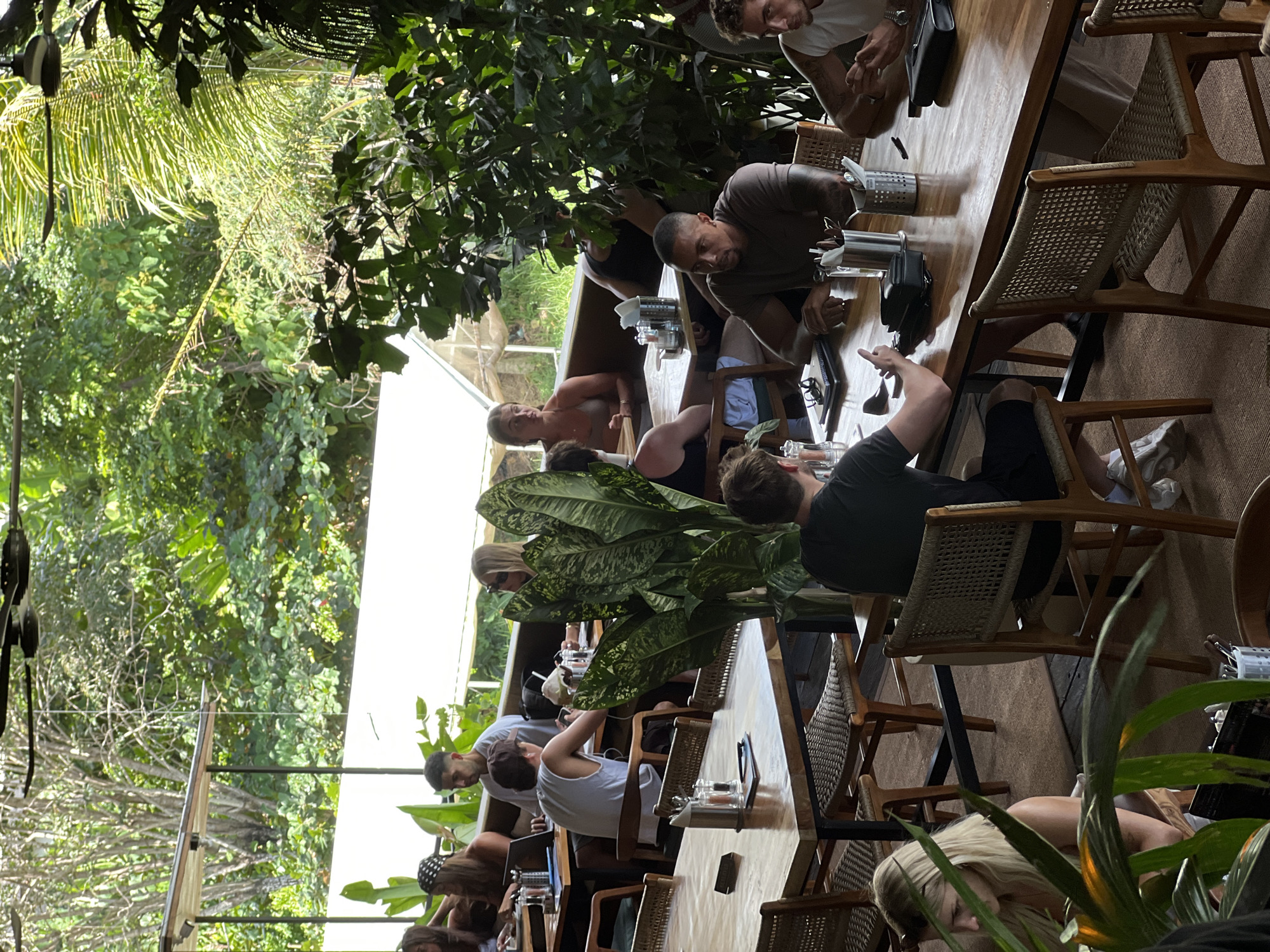 The image depicts an outdoor dining area of a restaurant in Mengwi, Indonesia, characterized by lush tropical foliage and open-air seating. Several groups of people are seated at wooden tables, engaged in various activities such as dining, conversing, and working on laptops.
In the foreground, a man with dark hair and a green t-shirt is seated at a table, gesturing with his left hand. To his right, a man with dark skin and a brown t-shirt is looking to his left, with his hands resting on the table. To the far right, a man with light hair and a white tank top is partially visible, looking down.
In the midground, a large, leafy plant with variegated leaves stands prominently on a table. A man in a grey tank top and a baseball cap is seated at the same table, looking down at his phone. To his left, a man in a white tank top is seated, with his back to the camera. Further left, a woman with blonde hair and a white top is visible, looking down.
In the background, more diners are seated at tables. A woman with blonde hair and a black and white patterned headband is looking at a laptop. To her right, a man in a grey t-shirt is seated, with a laptop in front of him. A woman with blonde hair and sunglasses is visible to the right, looking towards the left. Behind the tables, a white canopy structure and dense green foliage are visible, typical of the tropical environment of Mengwi. The overall atmosphere appears to be relaxed and casual, with natural light filtering through the leaves.