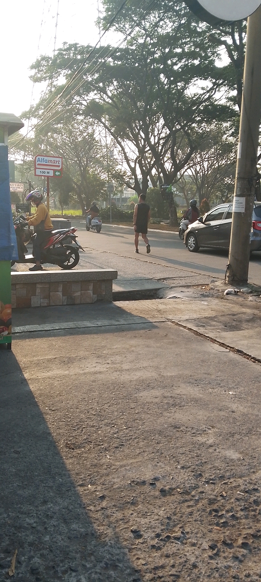 In Satriamekar, Indonesia, the image captures a sunlit street scene with a sidewalk in the foreground. To the left, a person wearing a yellow shirt and helmet sits on a red motorcycle. Across the street, a person in a black tank top and shorts walks away from the camera. Further down the road, another person rides a scooter, and a dark-colored car drives past. A sign indicating "Alfamart 100 M" with an arrow points to the right. The background is dominated by the lush foliage of large trees and overhead power lines. The ground is paved with asphalt and concrete, showing some cracks and debris. The overall impression is that of an ordinary day in a suburban or commercial area with moderate traffic and pedestrian activity.