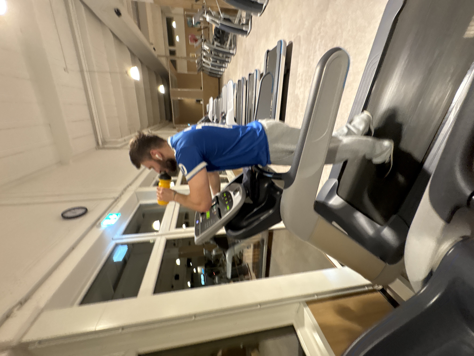 A man in a blue jersey and gray sweatpants stands on a treadmill holding a yellow water bottle in a gym. Other treadmills are visible in the background. The location appears to be in Mönchengladbach, Germany, but further information to confirm the city is not visible.