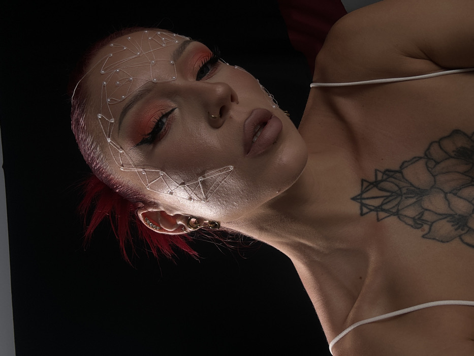 In Dalung, Indonesia, a close-up, studio-style portrait captures a female-presenting individual against a uniformly black background. The subject has red hair, styled upwards at the crown, and wears elaborate makeup featuring orange eyeshadow with black winged eyeliner and nude-toned lipstick. A small gold hoop nose ring is visible on the right nostril. A translucent, geometric facial adornment crafted from clear strands and small white beads covers portions of the forehead, temples, and cheekbones. The visible left ear displays multiple piercings, including small studs along the helix and a gold-colored chained earring in the lobe. The individual wears a white spaghetti-strap garment, revealing collarbones and a large black ink tattoo on the upper chest/sternum area, which combines geometric designs with floral patterns. The head is tilted downwards and slightly to the viewer's left, with eyes cast down and lips slightly parted. Front-left lighting illuminates the subject, creating highlights and shadows that define facial contours and the textured adornment.