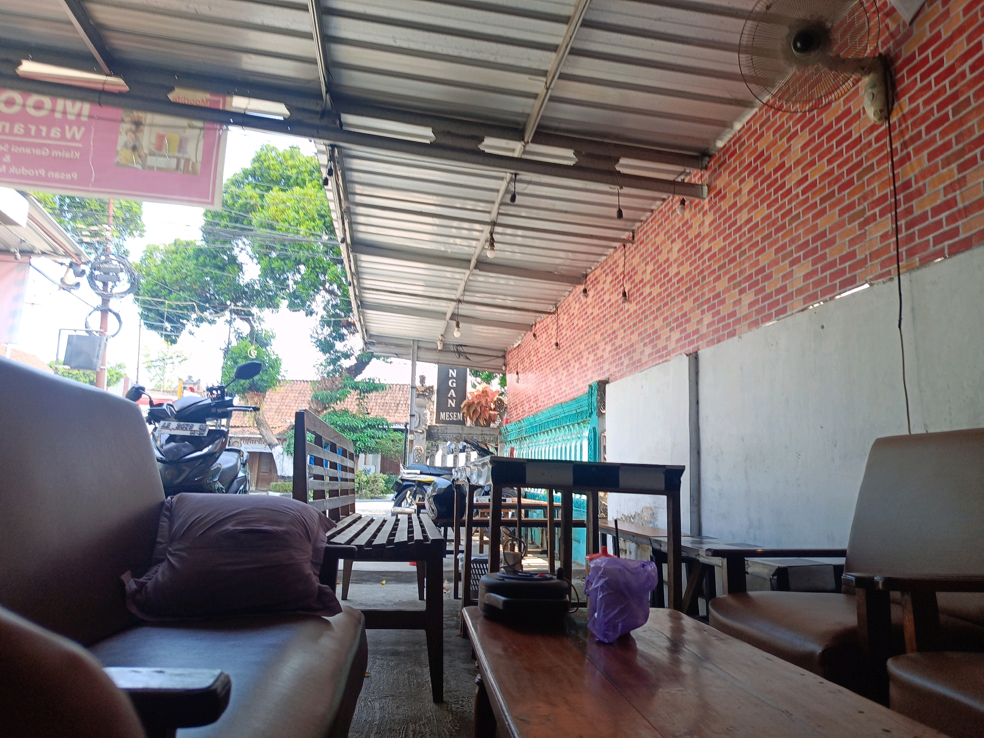 The image depicts an outdoor seating area, likely a cafe or restaurant, located in Candirejo, Indonesia. The scene is viewed from a low angle, focusing on a wooden table in the foreground with a purple plastic bag and a dark object on it. Behind this table, a brown leather couch is visible, with a dark-colored pillow resting on it. Further back, a wooden bench is situated. A motorcycle with a license plate is parked behind the bench. In the background, there are more tables and chairs, some with a checkered pattern. The seating area is covered by a corrugated metal roof supported by metal beams. A brick wall with a textured finish forms the right side of the structure. A wall-mounted fan is visible on the brick wall. Overhead, fluorescent lights are attached to the roof. Green trees and traditional Indonesian houses are visible beyond the seating area, suggesting a rural or semi-rural setting. A sign with Indonesian text is also present. The overall impression is of a relaxed, open-air environment.