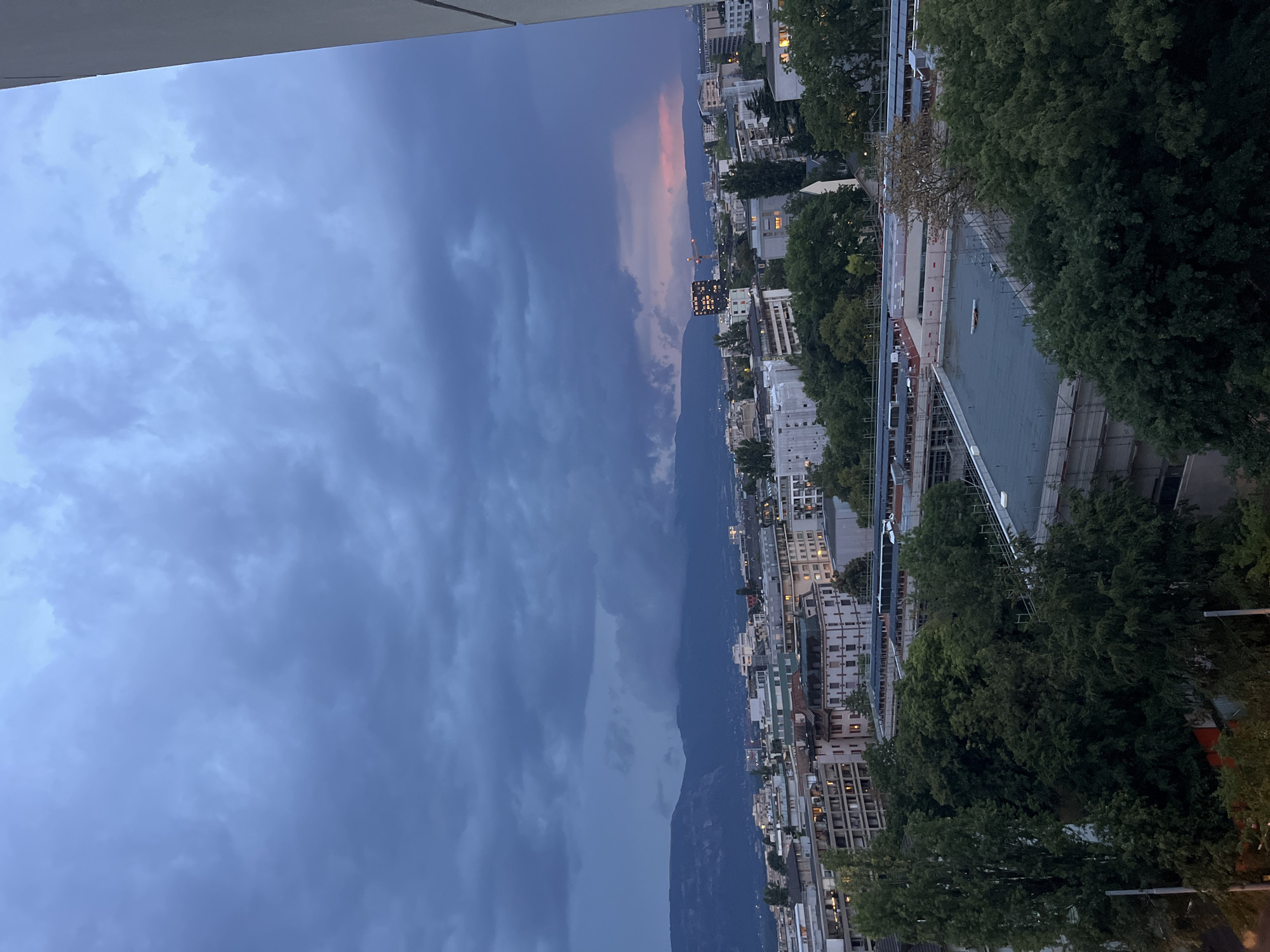 This image captures a panoramic view of Geneva, Switzerland, at dusk. The sky is dominated by a dramatic display of dark, heavy clouds, with a hint of pink and orange light peeking through on the horizon, suggesting the last moments of sunset or the approach of a storm. In the mid-ground, a dense urban landscape unfolds, characterized by a mix of residential and commercial buildings of varying heights and architectural styles. Many buildings display illuminated windows, indicating that some residents are at home. Green trees are interspersed throughout the cityscape, some reaching considerable heights and partially obscuring the lower levels of the buildings. In the background, rolling hills or mountains are visible, softened by the twilight and atmospheric haze. A sliver of a white building, likely a modern structure, is visible in the far right of the frame, partially cropped. The overall impression is one of a city transitioning from day to night under a moody and dynamic sky.