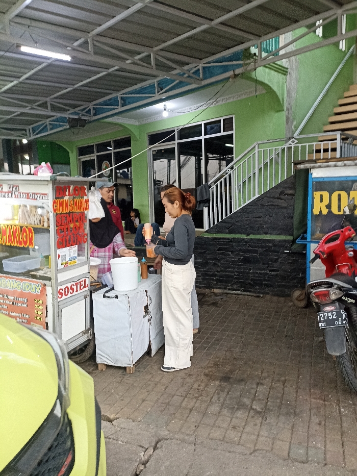 The scene is set in front of a small shop in Jatinangor, Indonesia, at the coordinates -6.935 latitude and 107.773 longitude. A woman in a white shirt and white pants is standing in front of a food cart, interacting with a woman in a black headscarf who is holding a white bucket. The woman in the white shirt is holding a glass. Behind them, a man in a red and white shirt is standing in front of the cart. There is a green car parked in the foreground, partially visible. To the right of the food cart is a red motorcycle with the number plate 2752. There is a set of stairs with a metal railing leading up to a building. The building has a green wall and large windows. The scene is outdoors, with a tiled floor and a metal roof overhead. There is a sign on the food cart with red and yellow text that says "BLOR CIMIN & MAKLO."