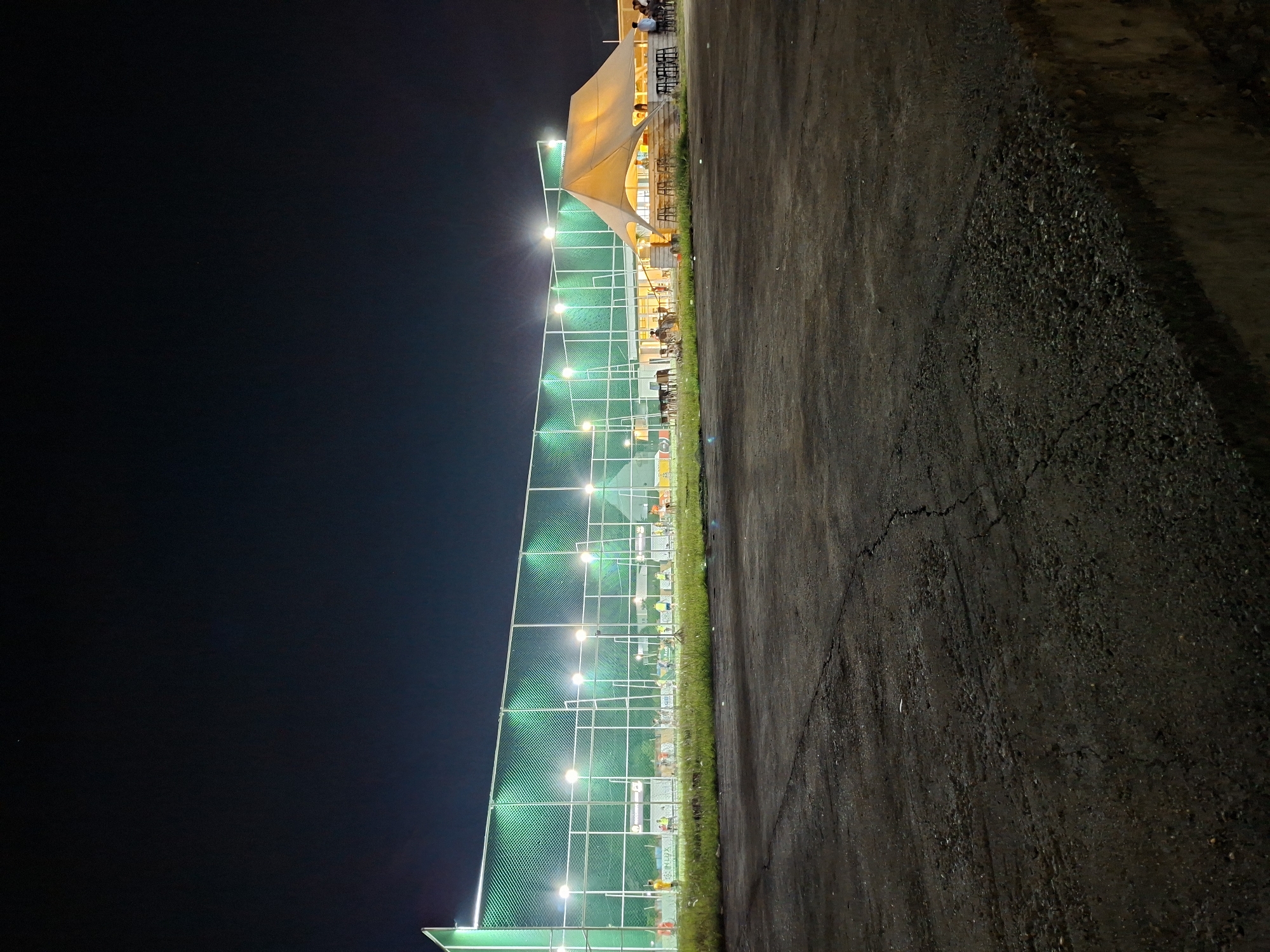 The image depicts an outdoor recreational complex at night, viewed horizontally. To the left, a sports court, possibly for futsal or badminton, is brightly illuminated by multiple floodlights. These lights cast a distinct greenish glow on the tall perimeter netting that encloses the court. A narrow strip of vibrant green grass runs along the edge of the court, separating it from a dark, rough-textured asphalt or concrete path, which occupies the foreground on the right. This path shows visible cracks.
Adjacent to the sports court, on the right side of the image, is a building with an expansive outdoor seating area. This area is covered by a large, light-toned, sail-like canopy, suggesting a cafe or restaurant. Underneath the canopy, several people are visible, seated at tables and chairs, engaged in socializing or dining. The canopy area is illuminated with warm-toned lighting, creating a contrast with the cooler, brighter lights of the sports court.
The dark sky confirms the scene is set at night, under clear weather conditions. While no active game is underway on the court, its floodlit state indicates it is ready for use. Along the base of the court's fencing, there are several advertising panels; one indistinct logo or text might read "INARA". The overall atmosphere is that of an active commercial leisure facility in Lhokseumawe, Indonesia, providing options for both sporting activities and casual dining or gathering.