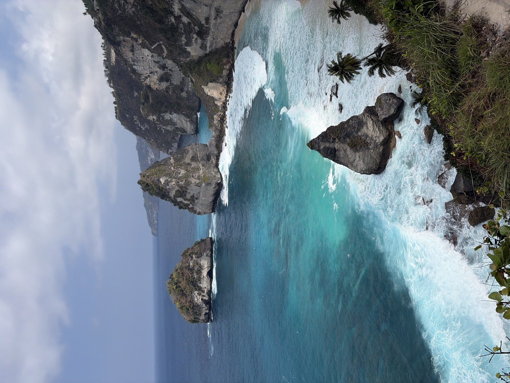 This is a high-angle, eye-level, medium shot of a dramatic coastal landscape in Nusa Penida, Indonesia. The scene is dominated by a rugged, cliff-lined coastline and a turquoise ocean. Several large, weathered rock formations rise from the sea, some capped with vegetation. The water exhibits a striking gradient of blues, transitioning from deep navy offshore to vibrant cyan closer to the shore, where white waves crash against the rocks and a sandy beach. Lush green vegetation and palm trees cling to the steep cliffsides, indicating a tropical environment. The sky above is partly cloudy with a soft, diffused light. There are no people or animals visible in the image. The overall impression is one of natural beauty and raw coastal power.