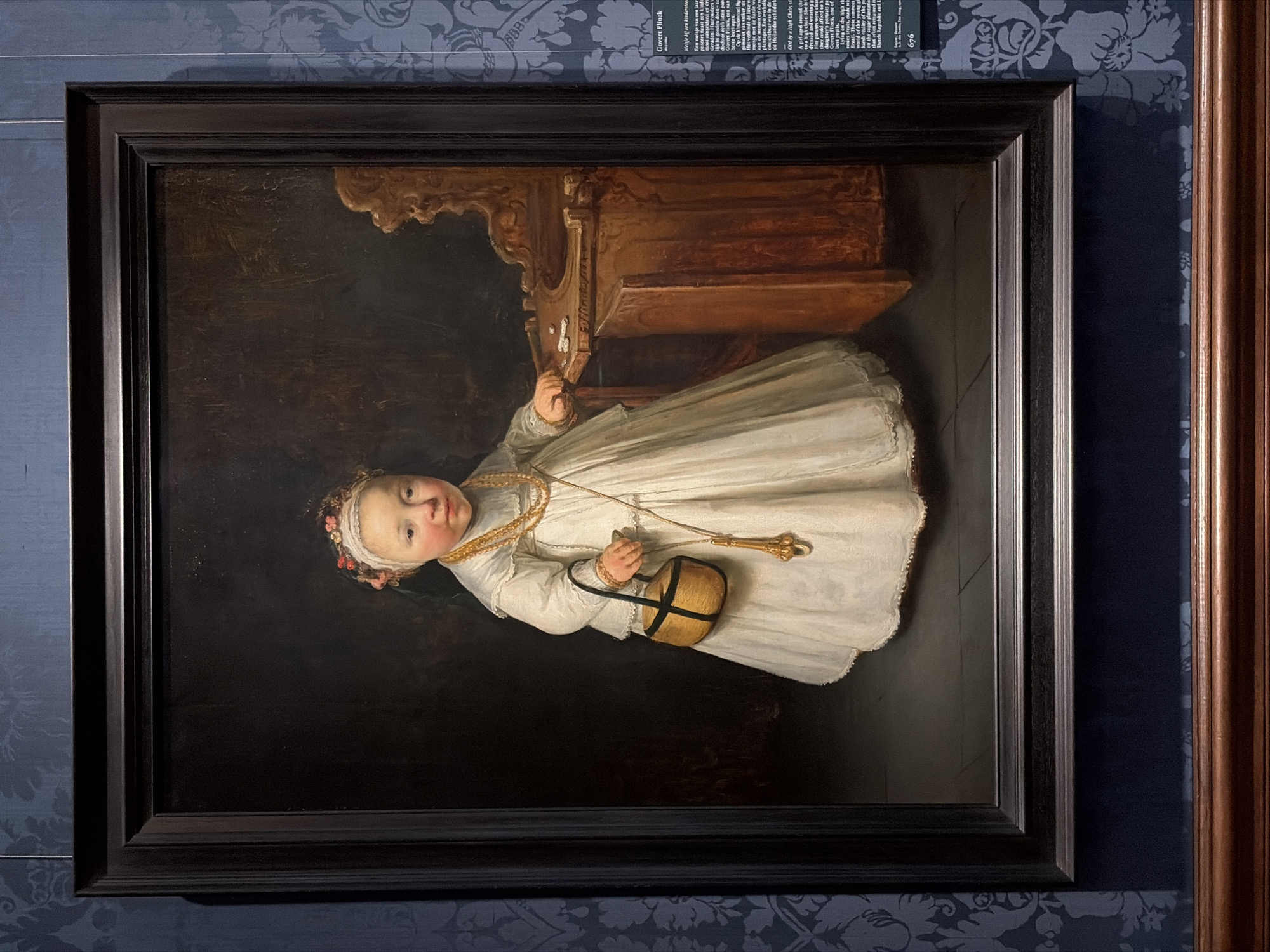 The image depicts a painting titled "Girl by a High Chair" by Govert Flinck, hung on a wall in The Hague, Netherlands. The painting is framed in a dark, tiered wooden frame and shows a young child standing in front of a carved wooden high chair. The child wears a long white dress with gold jewelry and holds a small woven basket. The painting is hung on a wall with blue patterned wallpaper, and a portion of another wooden frame is visible to the right of the painting. A placard with descriptive text is visible above the painting.