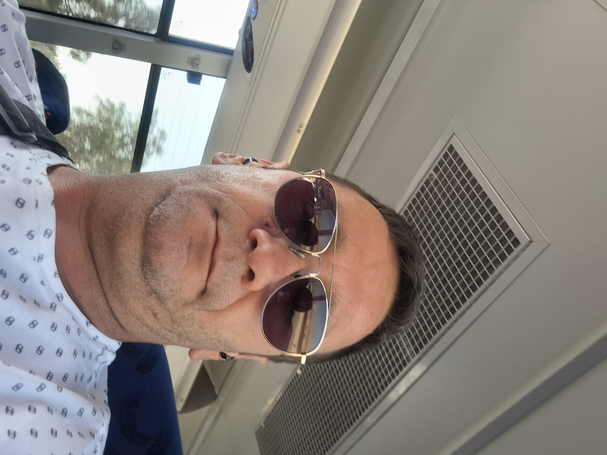 The image is a selfie taken by a man wearing sunglasses and a patterned white shirt. He has short, dark hair and stubble on his face. The man is inside a vehicle, likely a bus, as indicated by the ceiling panel with a grate and the windows with visible greenery outside. There are also seats visible in the background. The reflection in the sunglasses shows a landscape, possibly a road and trees. The man appears to be looking at the camera. The location is Bnei Shimon Regional Council, Israel.