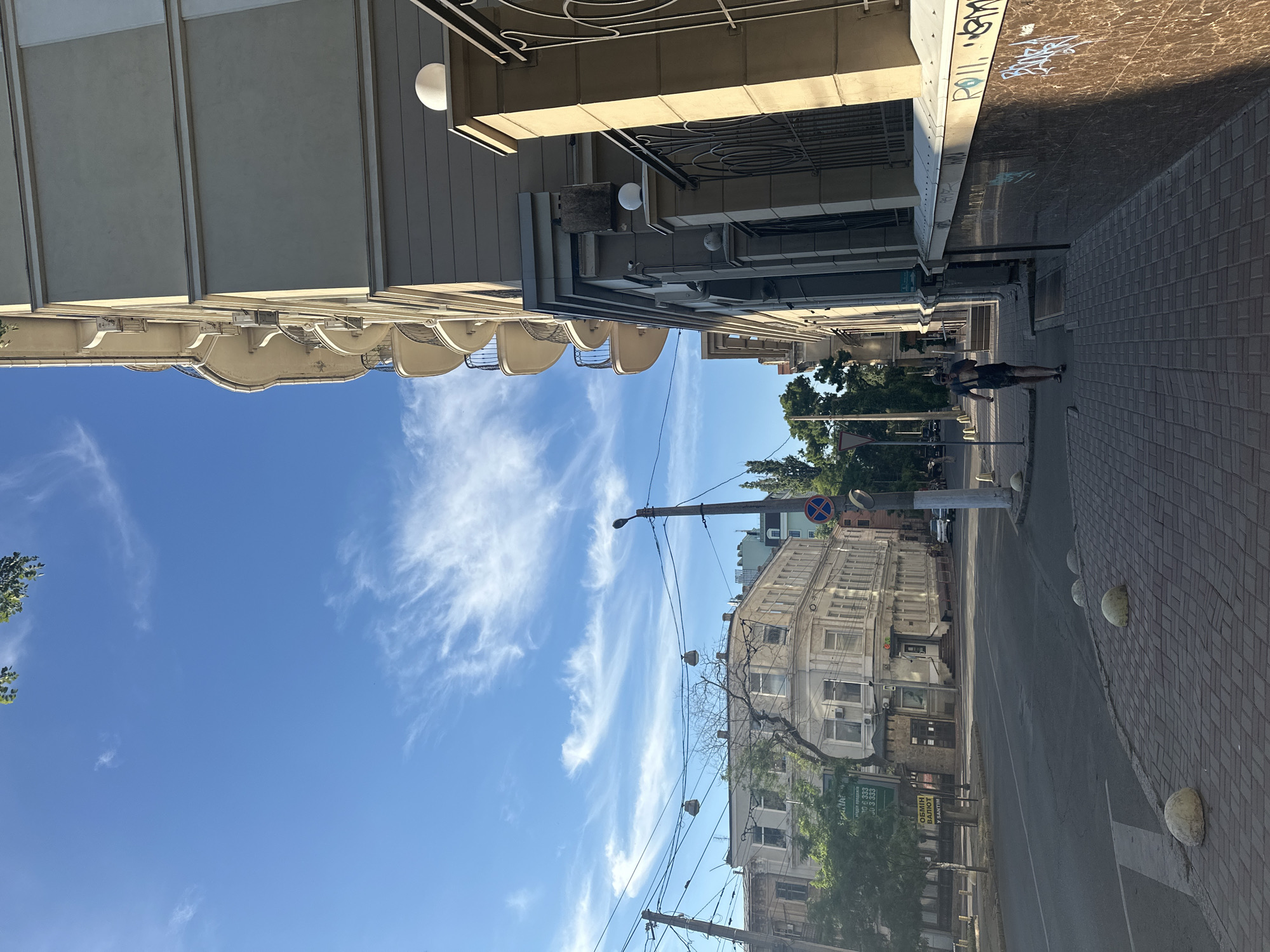 This is a vertical, full-shot photograph taken from a street level perspective looking upwards towards the sky and a street scene. The image is oriented sideways, with the sky occupying the left half and the buildings and street on the right.
The sky is a vibrant blue with thin, wispy white clouds scattered across it, suggesting a clear and sunny day. The sun is likely high in the sky, casting shadows and illuminating the scene.
On the right side of the image, a street runs diagonally away from the viewer. The street is paved with asphalt and bordered by a brick sidewalk. Several round, concrete bollards are placed along the edge of the sidewalk. A lone person, dressed in dark clothing, is walking on the sidewalk.
Buildings line the street. The closest building on the right is a multi-story structure with a curved facade, featuring many windows. Further down the street, other buildings of varying architectural styles are visible, some appearing older with ornate details, and others more modern. Trees with green foliage are interspersed between the buildings.
There are several streetlights and utility poles with numerous wires stretching across the scene. A traffic sign with a blue circle and a red border is mounted on a pole.
Graffiti with stylized lettering is visible on a wall above the sidewalk on the right side of the image. A green banner with text in a Cyrillic alphabet is also attached to a building. The text includes what appears to be a phone number with multiple instances of "333".
The overall impression is a typical urban street scene in daylight, likely in a European city. The atmosphere appears calm and ordinary.