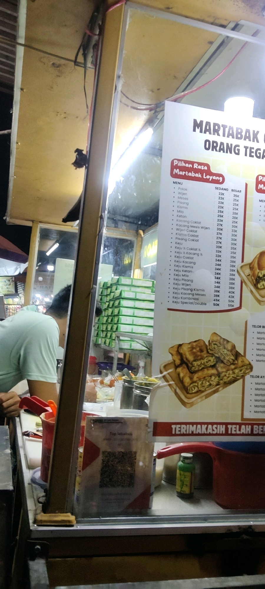 In Cibatu, Indonesia, a food stall vendor, a man wearing a light blue shirt, is seen from behind, working at a counter. The stall has a yellow exterior and is illuminated by artificial lights. A menu board is prominently displayed, featuring text in Indonesian. The menu lists various flavors of "Martabak Loyang," a type of pancake, with prices indicated in Indonesian Rupiah (K). The items on the menu include "Polos," "Wijen," "Meses," "Pisang," "Kismis," "Milo," "Ketan," and several cheese-based variations like "Keju Coklat," "Keju Kacang," and "Keju Kombinasi." Beside the menu, an illustration depicts several pieces of cooked martabak on a wooden board. In the foreground, a QR code is visible on a stand, likely for payment or information. Behind the vendor, stacks of green boxes are visible, possibly containing food packaging or supplies. A red plastic container and a small green bottle are also present on the counter. The overall environment suggests a bustling street food scene at night.