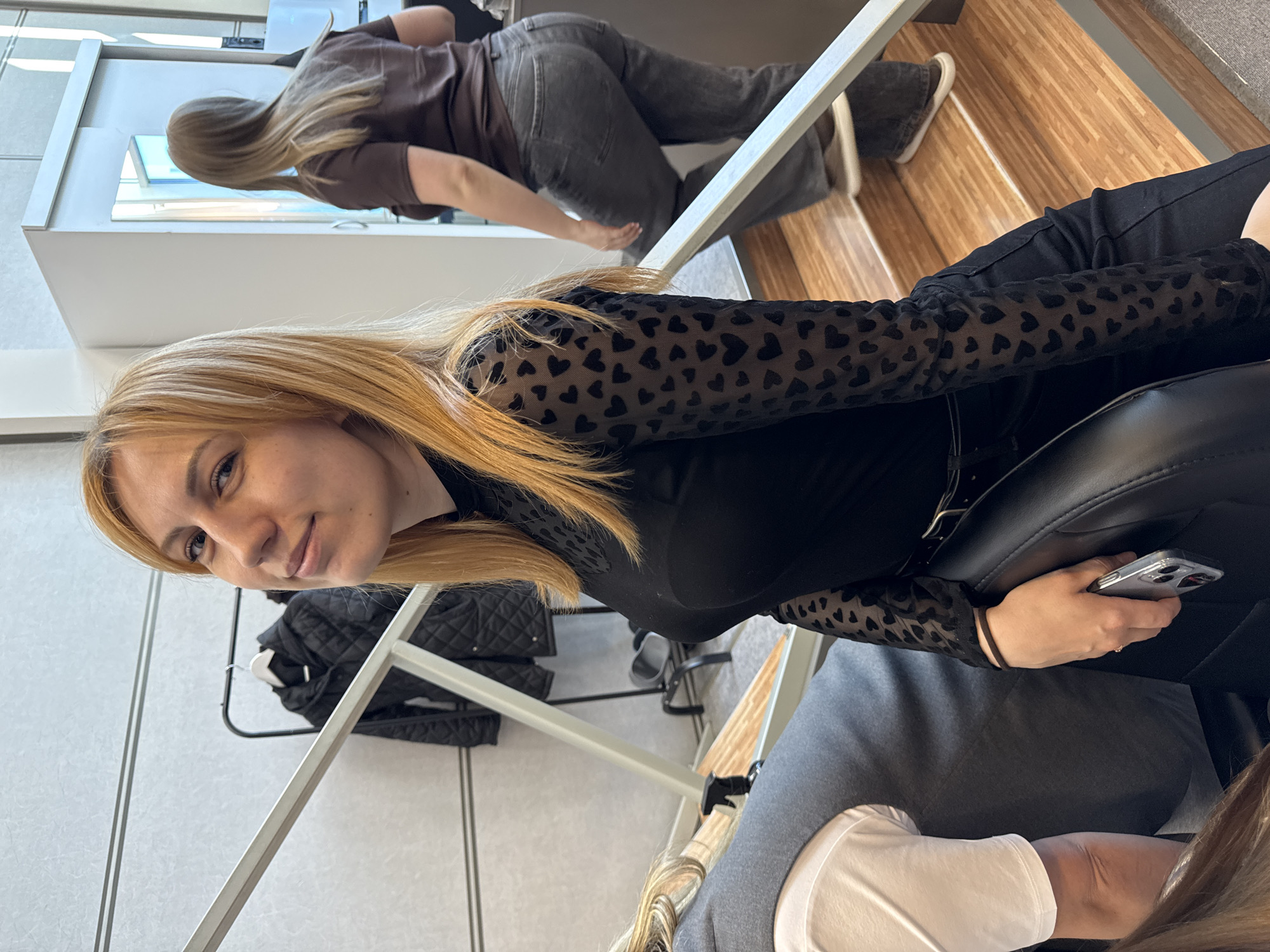Scene observed in Perm, Russia, depicting three females in an indoor setting. One fair-skinned female is in the foreground, seated and holding a cell phone. She is wearing a black top with a heart pattern on the sleeves. Behind her, a quilted black bag is visible near a white structure, possibly a staircase railing. Another female is partially visible in the upper portion of the frame, wearing a brown shirt and grey jeans, seemingly climbing stairs. A third female is partially visible in the lower right corner wearing white, with blonde hair. The environment includes a staircase with wooden treads, and a wall with light-colored, vertical paneling.