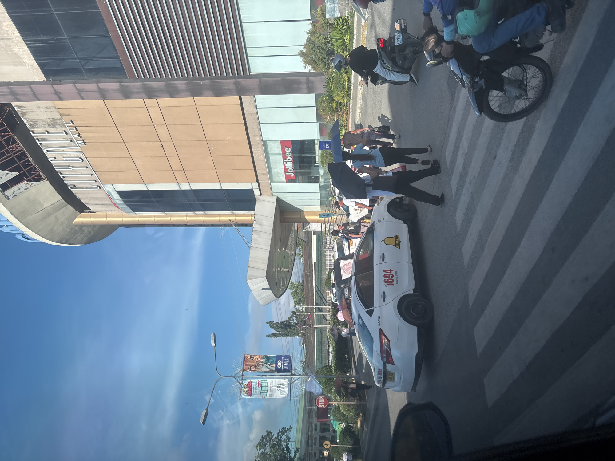 The image captures a street scene in front of the Citygate Mall in Davao City, Philippines. The mall building, with its distinctive tan facade and "CITYGATE MALL" signage, dominates the upper right portion of the frame. Below it, a Jollibee restaurant is visible.
In the foreground, a white taxi with the number "1694" and a bell logo on its side is stopped at a pedestrian crossing. The taxi has a license plate reading "AAG-1820". Several people are present on the street. Some are walking, one is holding a black umbrella, and another is partially obscured by the taxi. Two motorcycles are also visible: one parked on the right side with a rider wearing a helmet, and another partially visible motorcycle in the lower right corner with a rider.
A street lamp with two lights and two banners hanging from it stands on the left side of the road, one banner advertising "BACK TO SCHOOL". A red "STOP" sign is also visible near the pedestrian crossing. The sky is bright blue with some scattered clouds. The overall environment suggests a busy urban setting with a mix of vehicles and pedestrians.