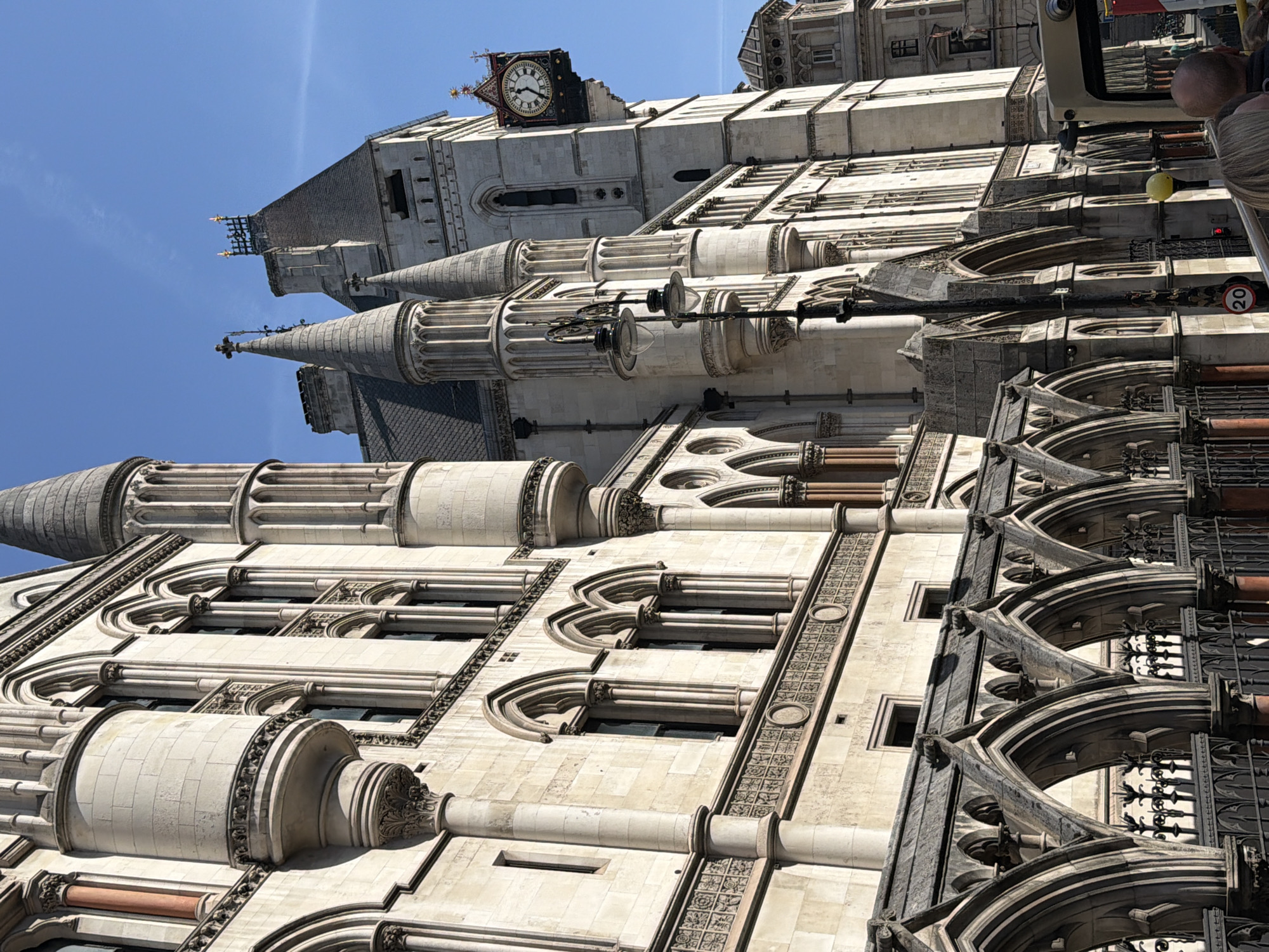 The image depicts the Royal Courts of Justice in London, United Kingdom. The building is constructed of light-colored stone and features a distinct Gothic Revival style. It showcases intricate architectural details, including arched windows, decorative moldings, spires, and turrets. The sky is clear and blue. The camera angle is tilted upwards, emphasizing the height and grandeur of the structure. A clock tower can be seen at the top of the building.