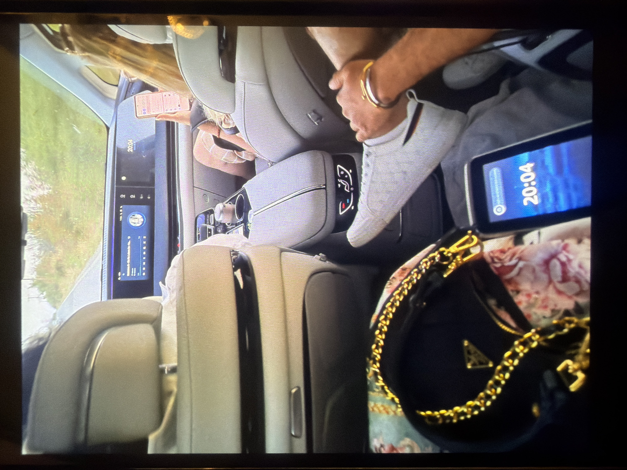The image captures the luxurious interior of a modern car, likely a sedan or SUV, from an elevated perspective looking towards the front. The setting appears to be during the evening, indicated by the time "20:04" visible on multiple screens.
In the foreground, a black handbag with prominent gold chain straps and a gold triangular "PRADA" logo rests on a floral-patterned fabric. To its left, a person's bare arm, adorned with a gold-colored bangle, is seen, with their foot, wearing a light grey sneaker, resting nearby.
Further into the vehicle's cabin, light-colored (possibly beige or light grey) leather seats and polished surfaces are visible. A central console features chrome accents and what appear to be touch-sensitive controls. Behind the front passenger, a person with blonde hair is engrossed in their smartphone, holding it with both hands. Their phone displays a grid of application icons on a light-colored background. Large digital screens are integrated into the front seatbacks and across the dashboard, some also displaying the time "20:04" and other interface elements.
Outside the car window, greenery suggests a natural or roadside environment. The overall scene depicts occupants in a high-end vehicle, possibly traveling, with a relaxed atmosphere. The time of day is 8:04 PM. The location is specified as Sofia, Bulgaria.