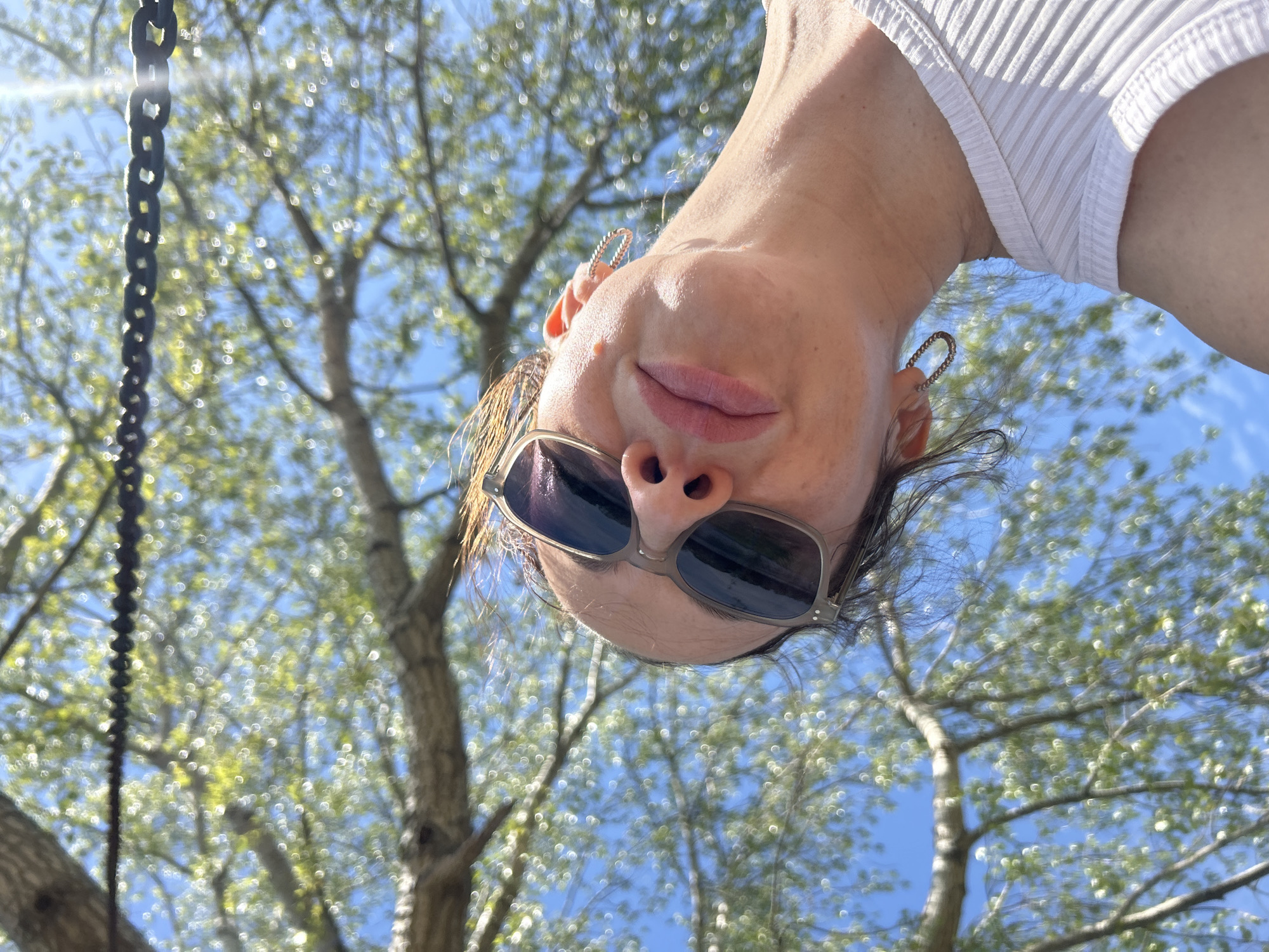 A low-angle shot depicts a person wearing sunglasses and earrings, with their face oriented upside down towards the camera. The background features a tree with light green foliage against a blue sky. A dark, possibly metal, chain is visible to the left. The person is wearing a white, ribbed top. The location is Chaikovsky, Russia.