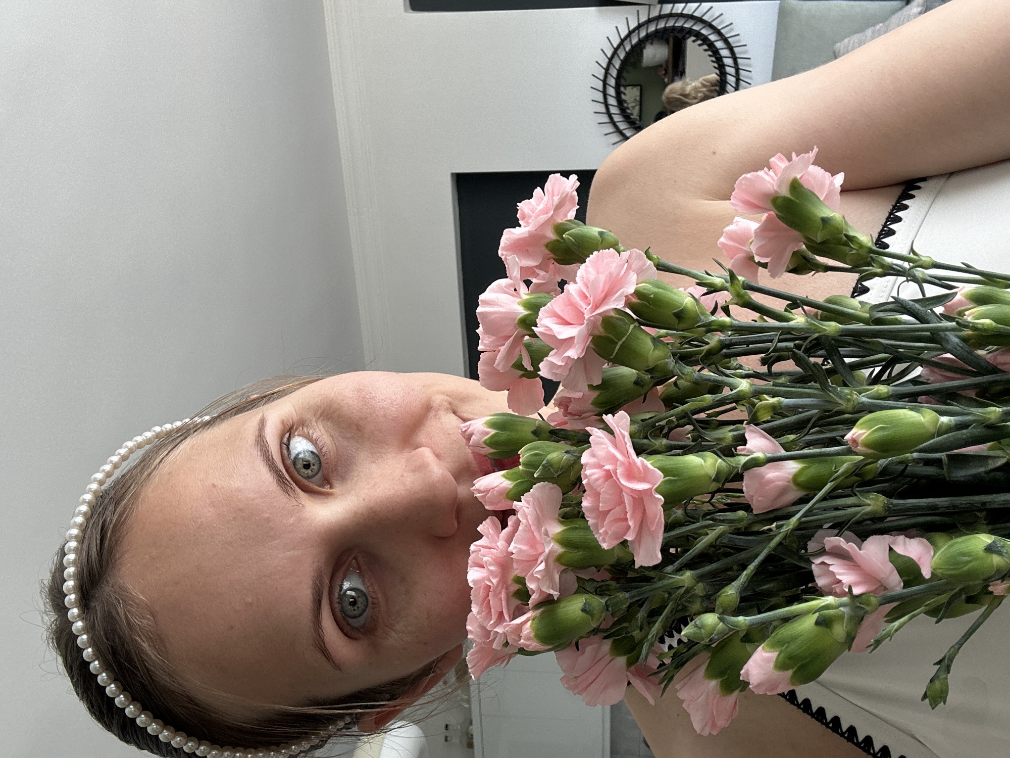 A woman is holding a bouquet of pink carnations close to her face. She is wearing a pearl headband. The background includes a wall, a dark rectangular object, and a circular decorative item with a black frame. There is no information to infer the location in Fethiye, Turkey.