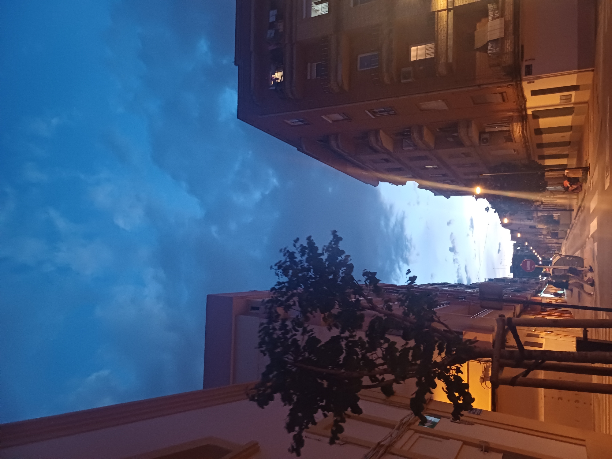 The image depicts a street scene in València, Spain, at dusk. Tall buildings line either side of the street, illuminated by streetlights and lights from within the buildings. The sky is visible above the buildings, displaying a mix of blue and dark clouds. A tree is visible in the foreground. People and vehicles can be seen further down the street.
