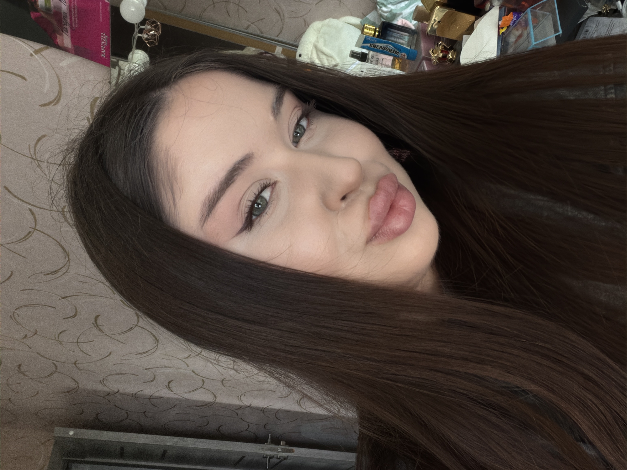 A close-up, waist-up, selfie-style photograph captures a young woman with long, dark brown hair and fair skin. She is facing the camera, her head tilted slightly to the right. Her eyes, with green irises, are open and looking towards the camera. She is wearing makeup, including winged eyeliner and eyeshadow, and her lips are full and slightly parted.
The background is out of focus but appears to be an indoor setting. To the left, a section of a door or cabinet is visible, with a textured wallpaper featuring a swirl pattern. To the right, shelves are visible with various objects, including bottles of perfume, cosmetic products, and decorative items. A pink and white package with the text "RAPUNZEL" is also visible on a shelf. The lighting in the scene is soft.