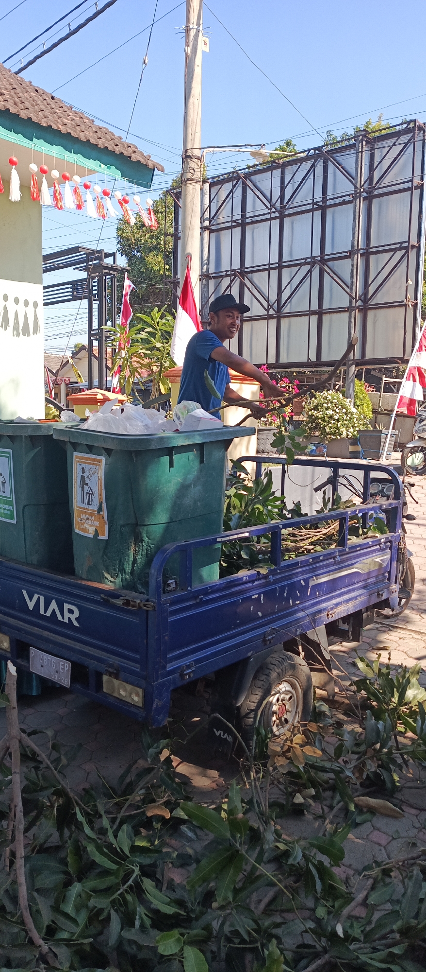 In the city of Arjasa, Indonesia, on a sunny day, a young man wearing a blue t-shirt and a black cap is standing by a blue three-wheeled vehicle. The vehicle has a large blue cargo bed filled with green trash bins and branches from plants. The man is holding some of the branches, appearing to be in the process of loading or unloading them. Two large green trash bins, one with a "Sampah Organik" (Organic Waste) label and a symbol of a person throwing trash, are visible in the cargo bed. The vehicle also has the brand name "VIAR" visible on its side, along with a license plate that reads "P 4976 EP 03.28". In the background, there is a large billboard structure and a building with red and white Indonesian flags hanging. The ground is paved with bricks, and fallen leaves and branches are scattered around the vehicle, suggesting recent yard work or debris collection. The overall scene depicts a routine waste management or yard cleanup activity in a public or semi-public outdoor space.