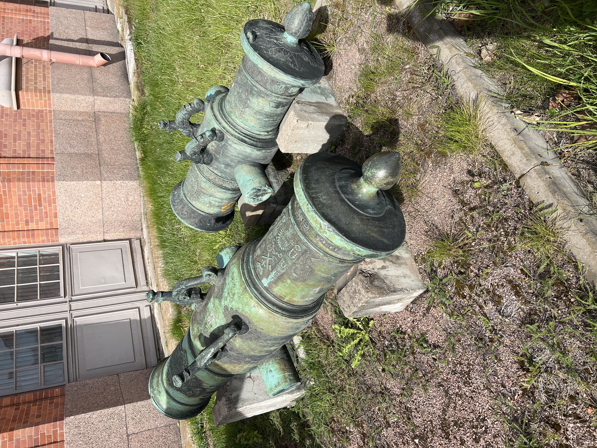 Two historic bronze mortars rest on stone blocks in an outdoor setting. Their green patina suggests age and exposure to the elements. The mortars are adorned with detailed carvings and designs, including what appears to be a finial atop each piece. In the background, there is a red brick building with white framed windows, indicating an urban location. The ground is covered in grass, gravel, and some weeds, suggesting the mortars are in a garden or park-like setting. Given the style and cultural significance, it is highly probable that the location is in the city of Saint Petersburg, Russia.